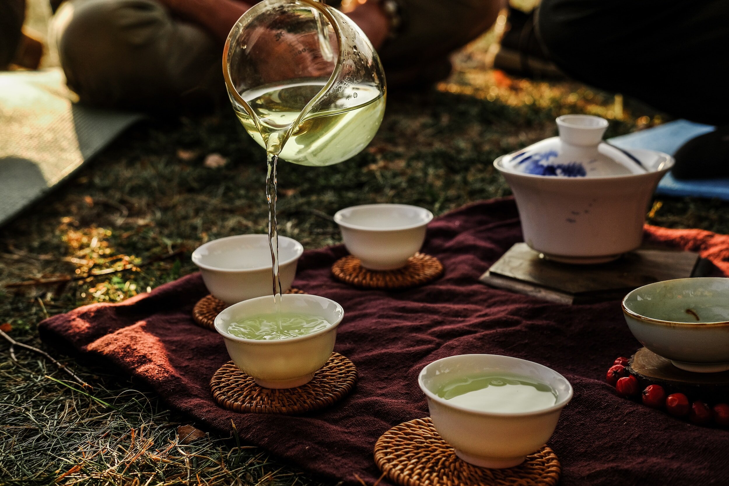 A tea ceremony setup with several small cups on a purple cloth, where a person is pouring green tea from a glass jug into one of the cups. The cups are placed on woven coasters and a larger tea pot sits nearby.