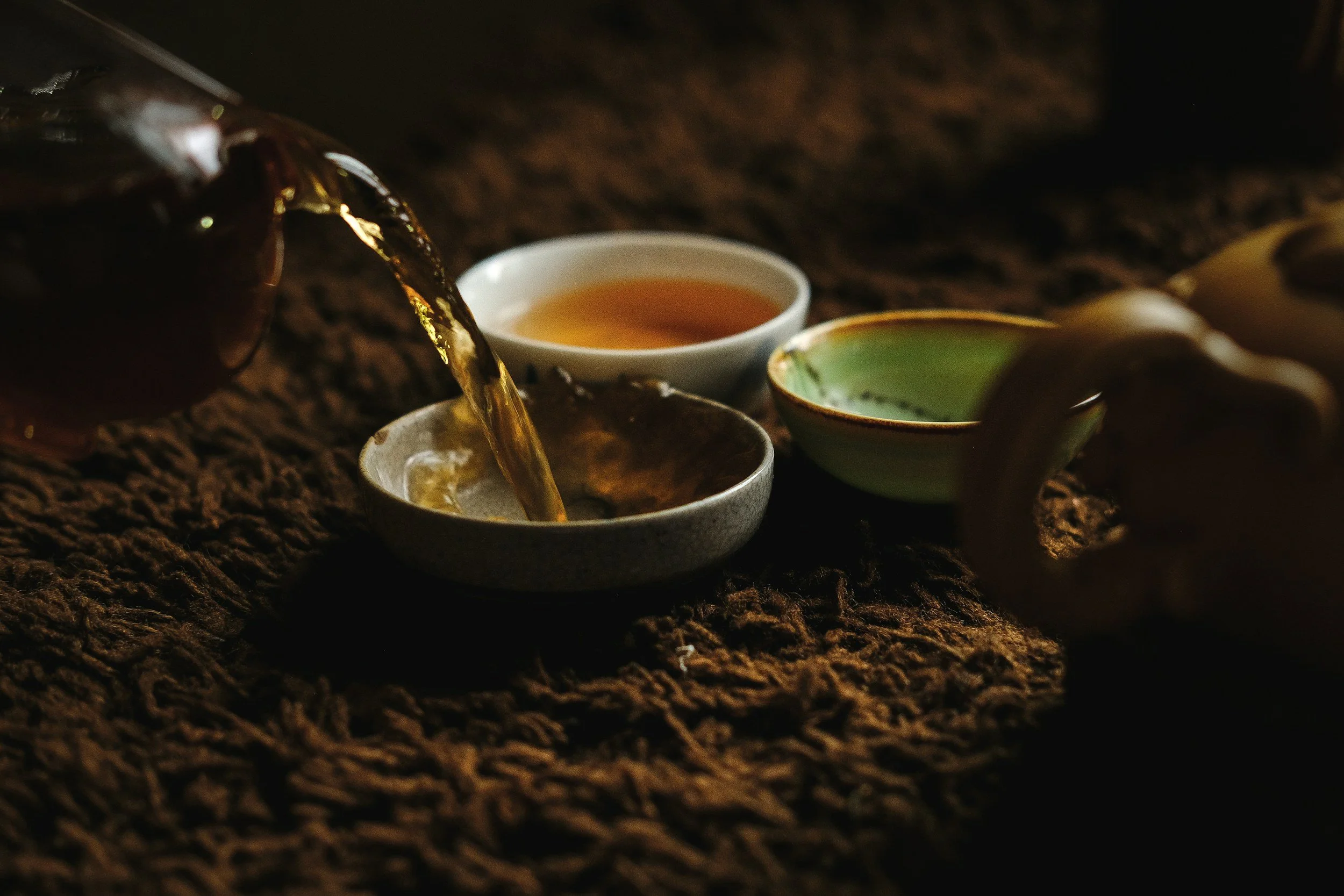 Tea being poured into a small bowl next to two ceramic cups on a textured surface.
