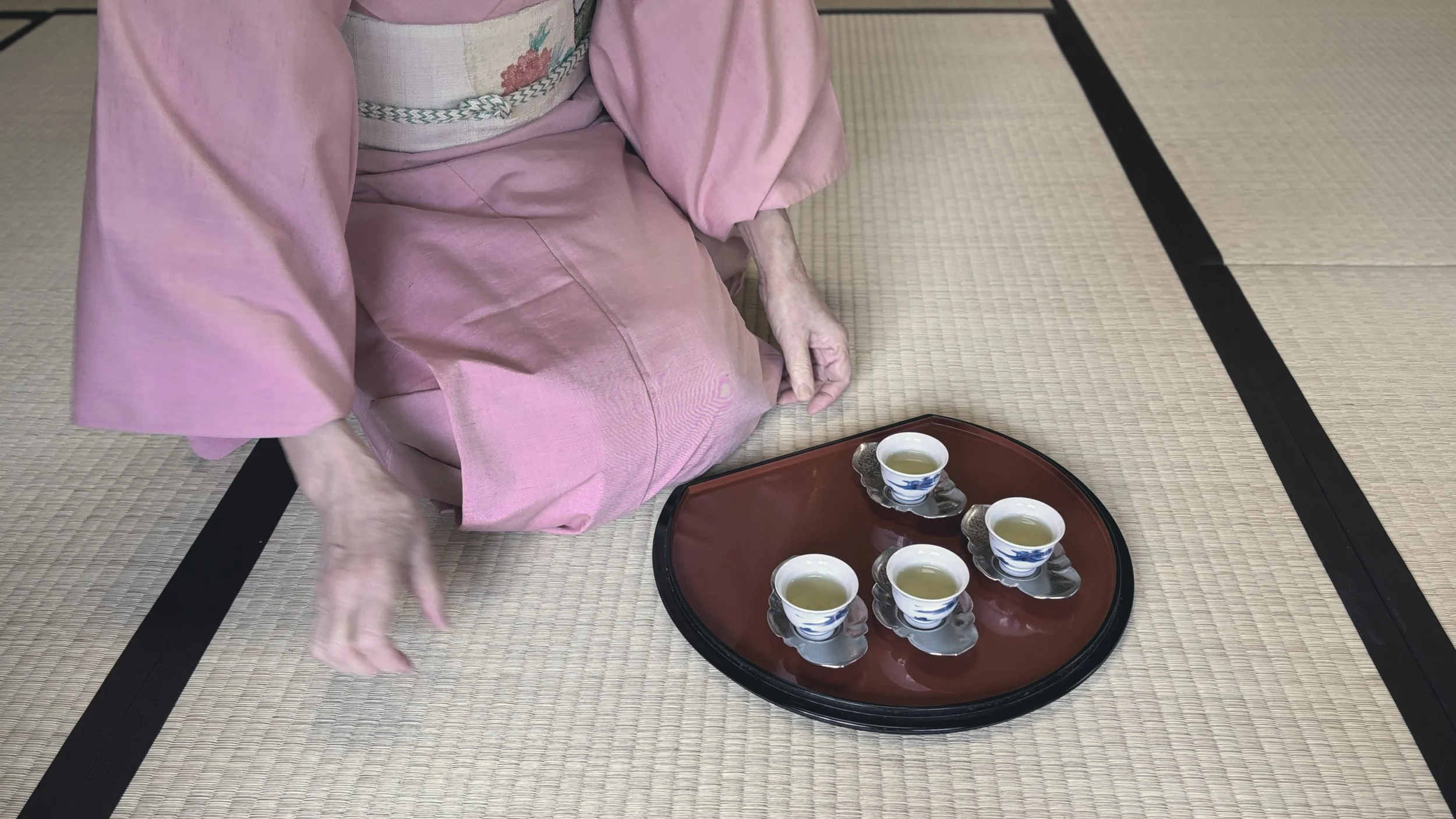 Person dressed in a pink kimono sitting on tatami mat floor, with a tray of five traditional Japanese tea cups in front of them, preparing to serve tea.