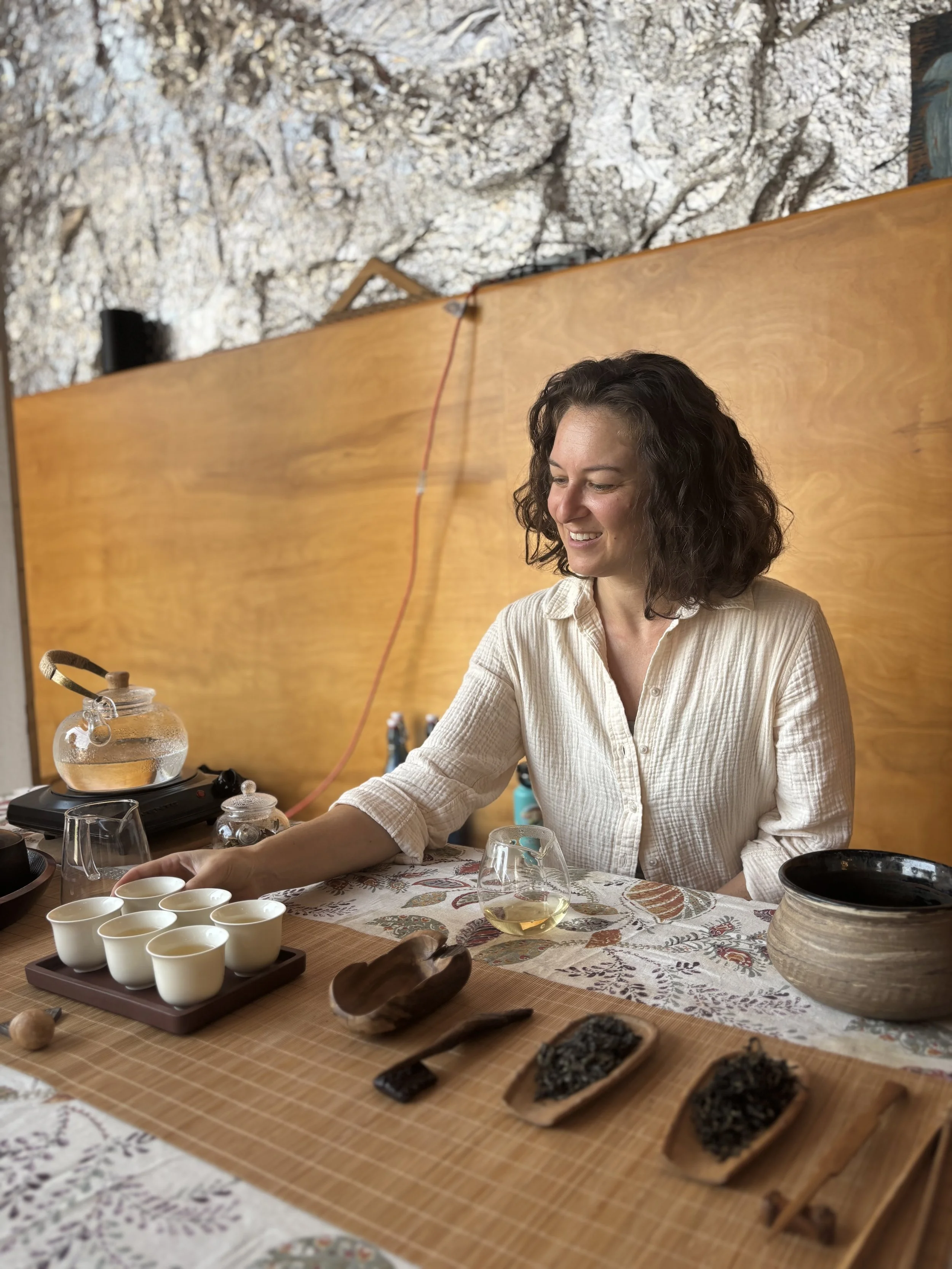 A woman with dark, curly hair sitting at a table with a tea set and various dried herbs and spices, with a rocky wall and wooden panel in the background.