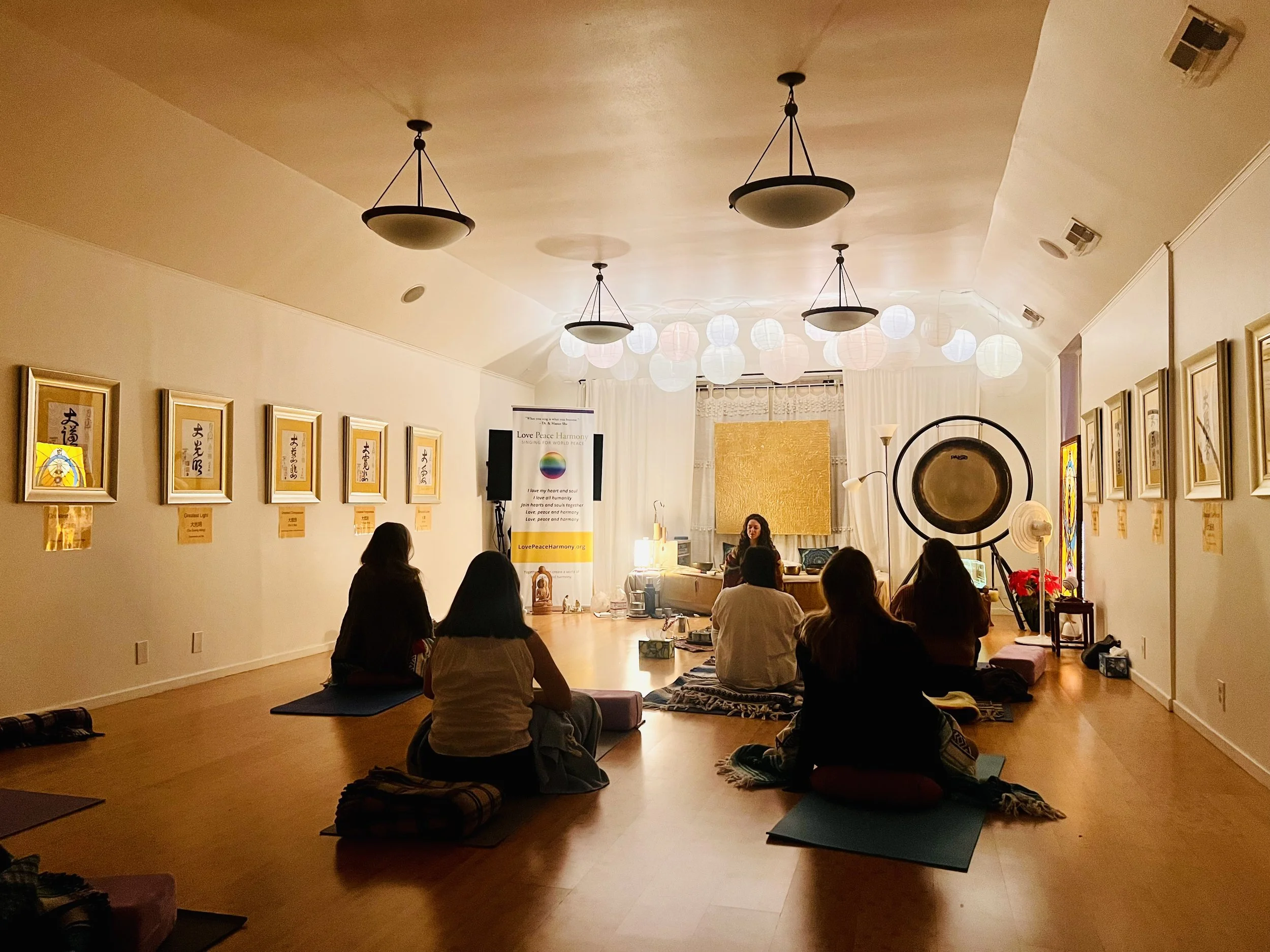 A group of people sitting on yoga mats or cushions on the floor in a room with warm lighting. The room is decorated with framed calligraphy art on one wall and paper lanterns hanging from the ceiling. A person is leading the session near a banner that reads "Love Peace Harmony."