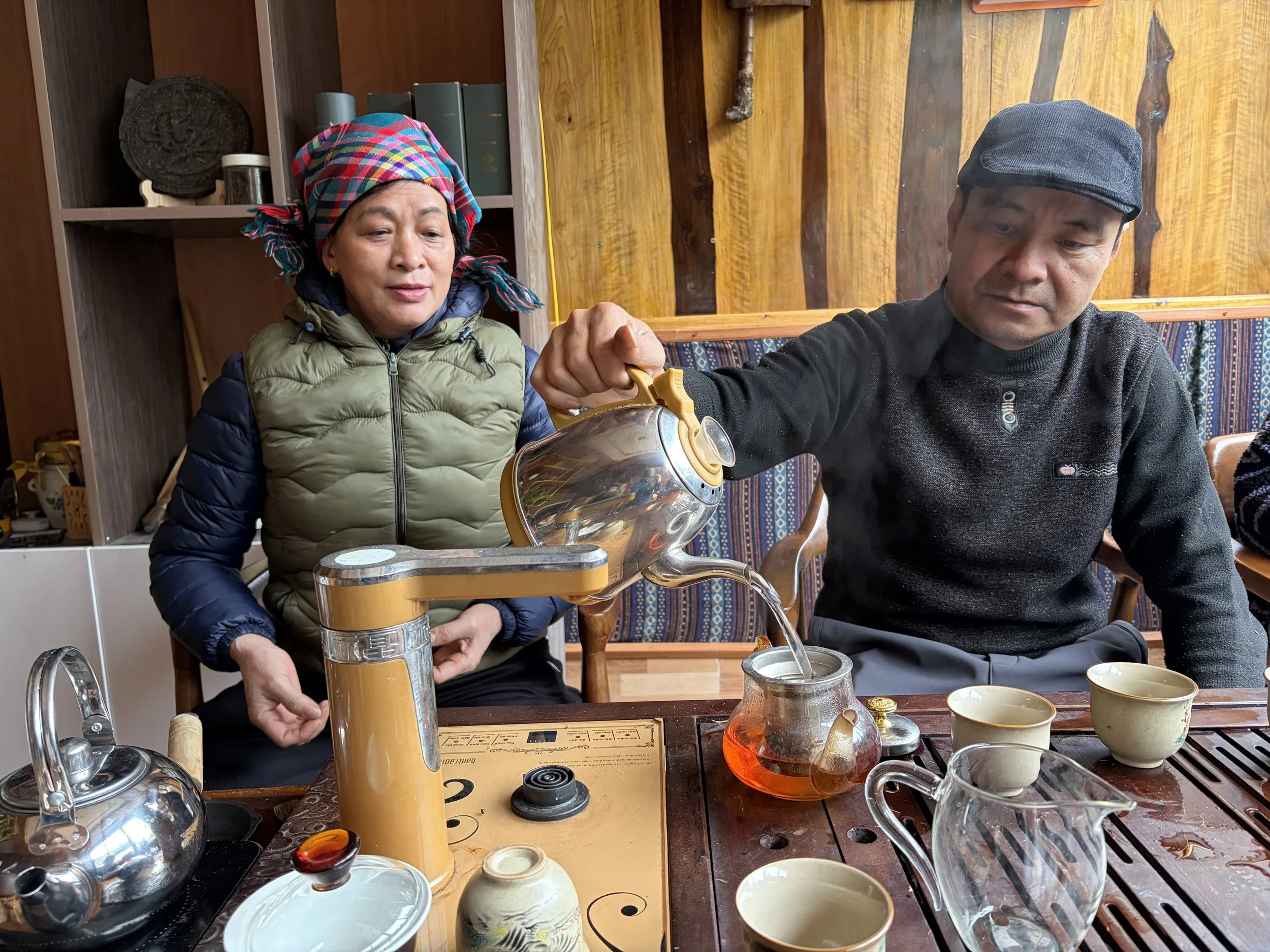 Two individuals sitting at a wooden table with tea cups and a teapot, preparing tea with a tea brewing device, in a cozy room with wooden walls and shelves.