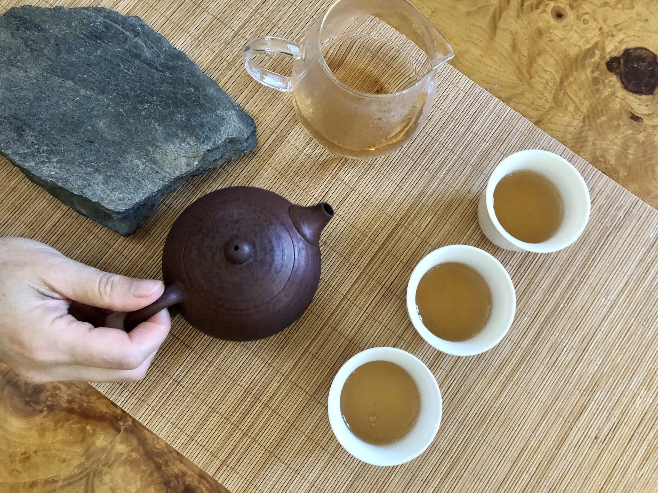 A person holding a small, round, dark brown teapot with a short spout. On a wooden table, there are three white cups filled with tea, a glass pitcher with tea, a dark stone slab, and a wooden item. The table has a bamboo placemat.