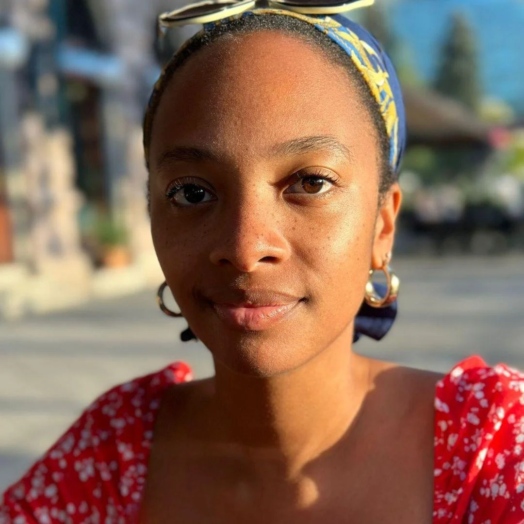 Close-up of a young woman with dark brown eyes and smooth skin wearing a yellow and blue patterned headband, gold hoop earrings, and a red top with white floral patterns, taken outdoors during golden hour.