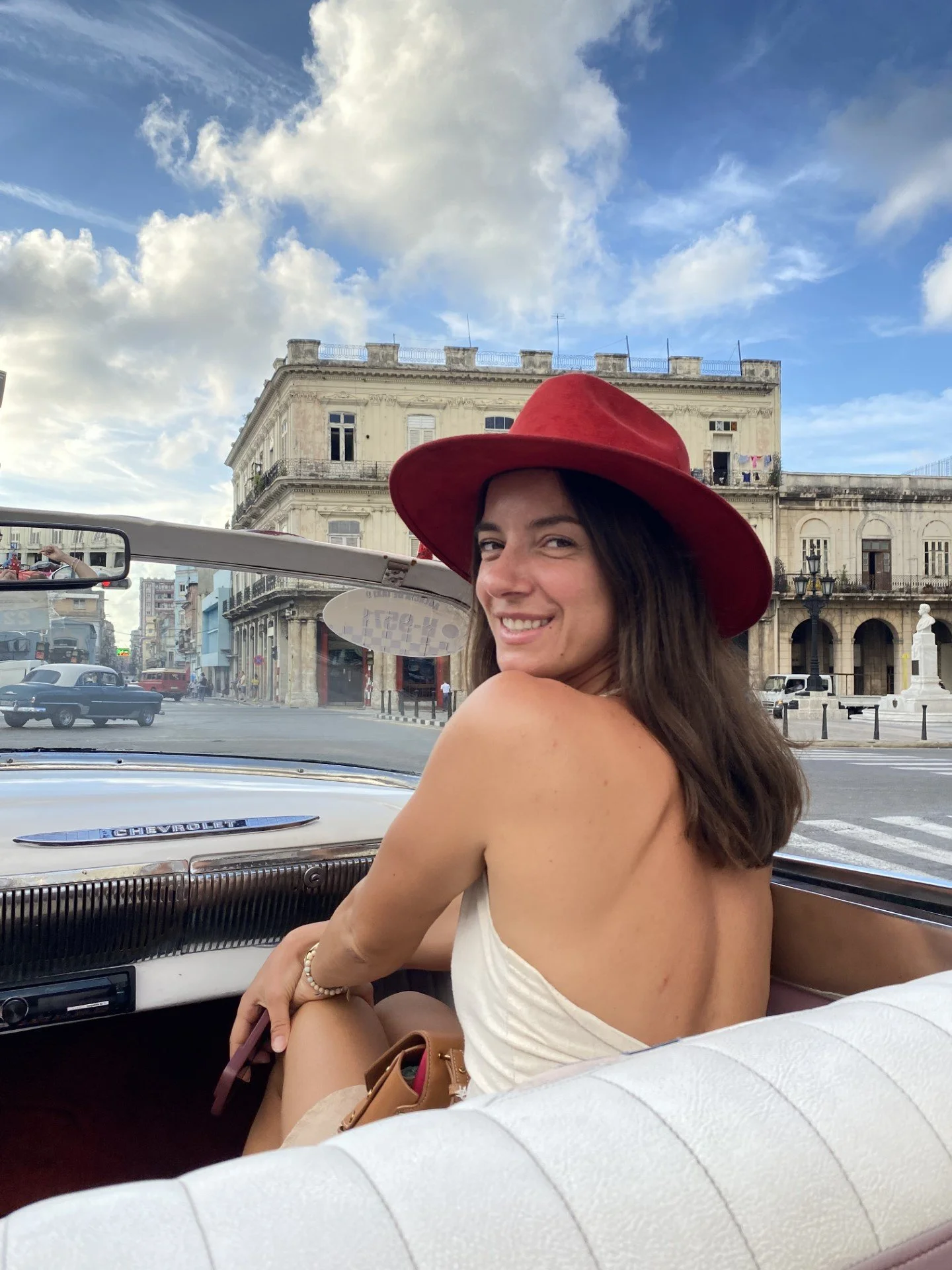 A smiling young woman wearing a red hat and white top, sitting in a vintage car, with an old building and blue sky with clouds in the background.
