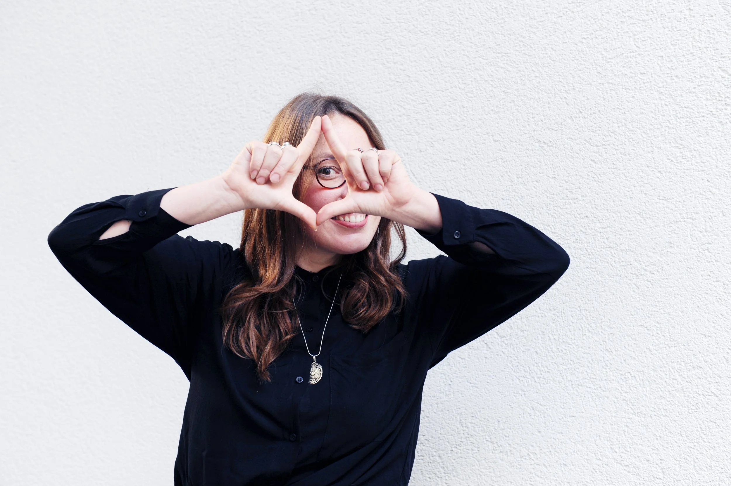 Frau mit braunen, gewellten Haaren trägt schwarze Bluse, macht ein Dreieck mit Händen vor Gesicht, lächelt, steht vor weißer Wand.