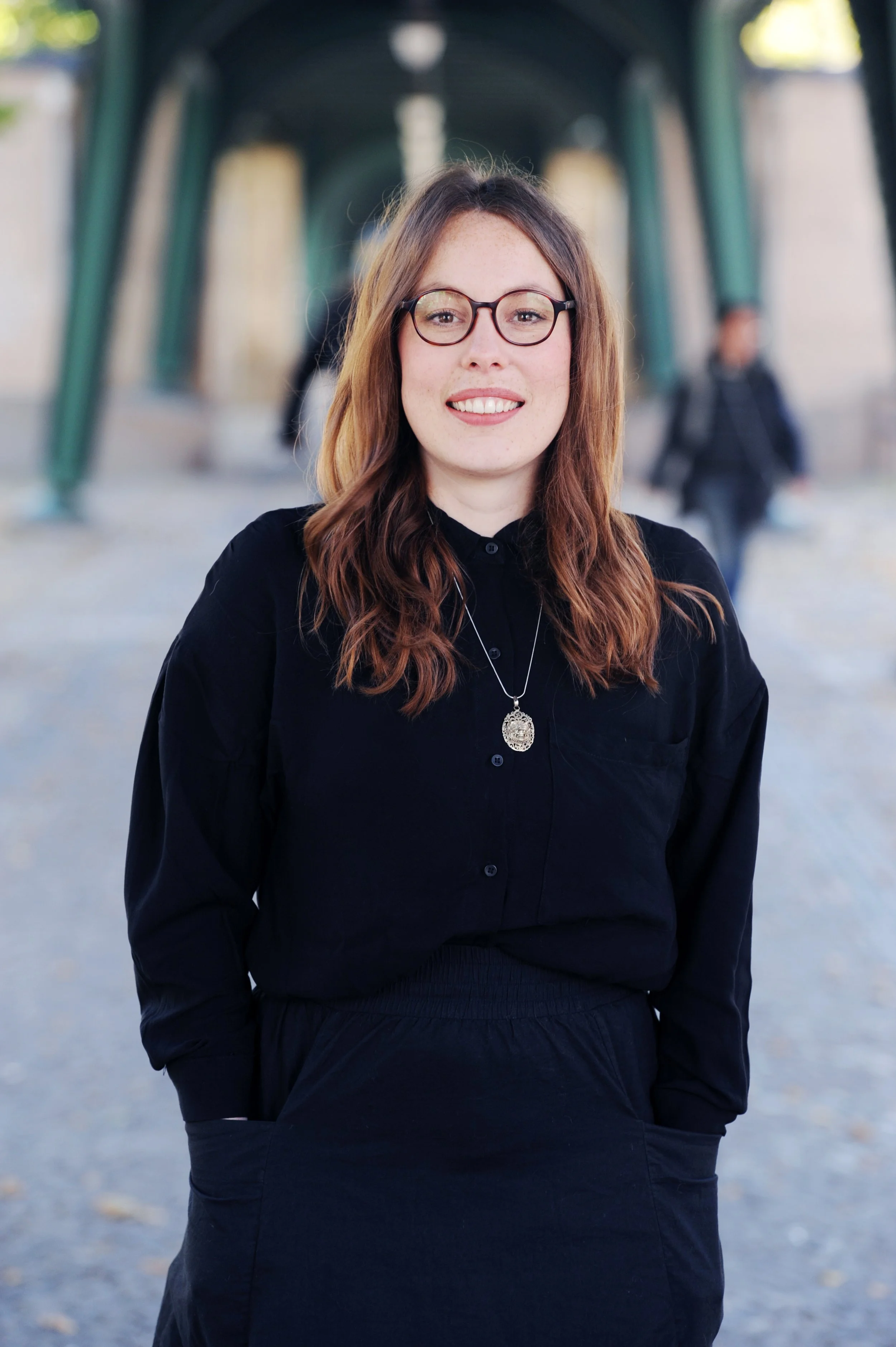 Eine lächelnde Frau - Elisa Schön - mit Brille und braunen, gewellten Haaren, die in einem schwarzen Outfit steht, im Hintergrund eine Brücke und Menschen.