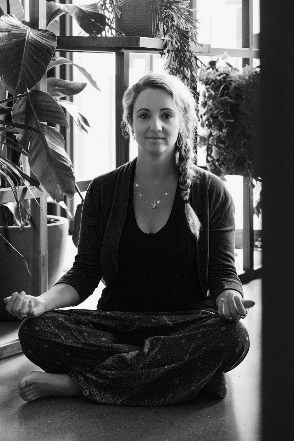 A woman practicing meditation in a room filled with large potted plants and sunlight coming through large windows, sitting cross-legged on the floor.