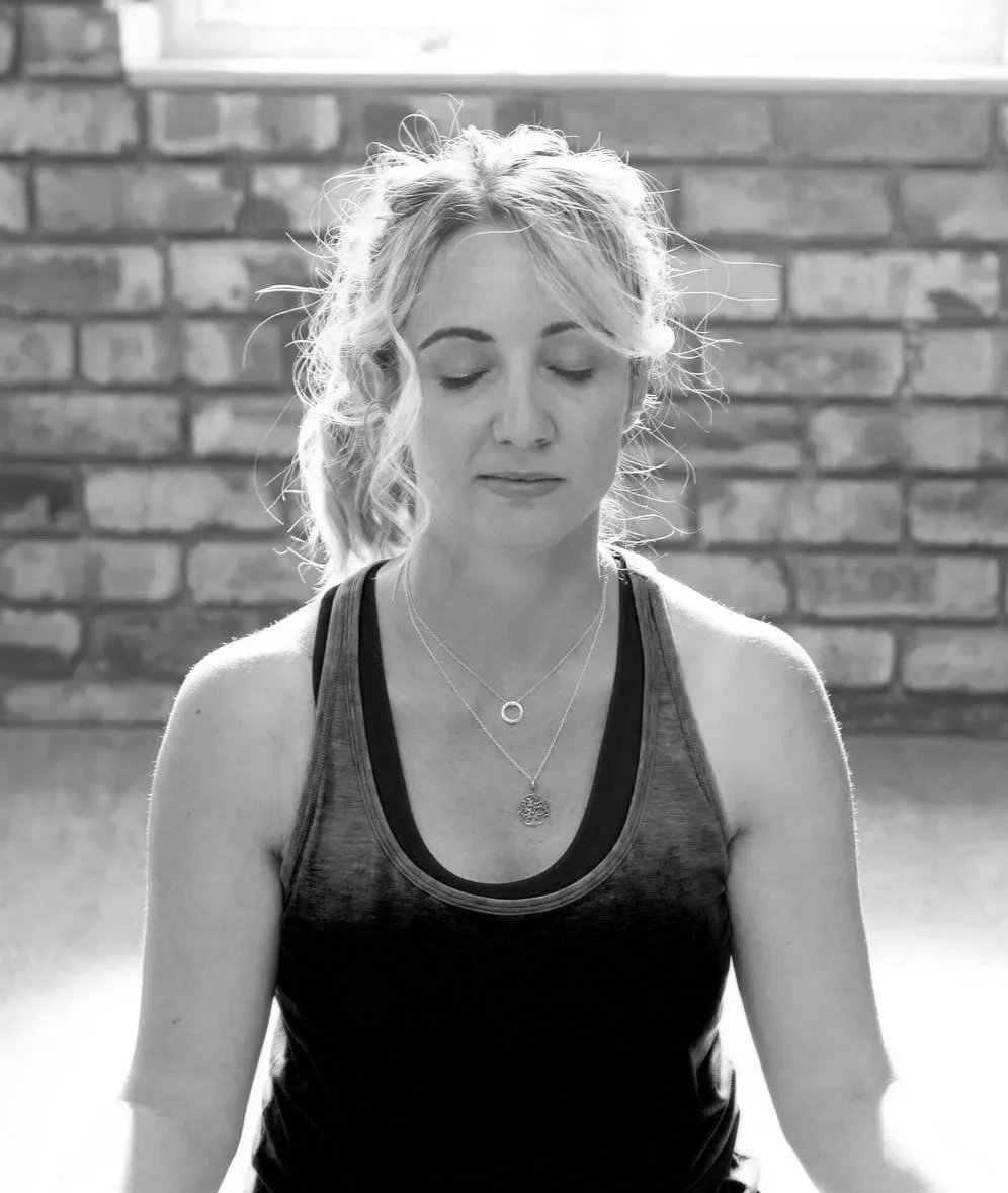 Black and white photo of a woman with curly hair in a ponytail, sitting with eyes closed against a brick wall background.