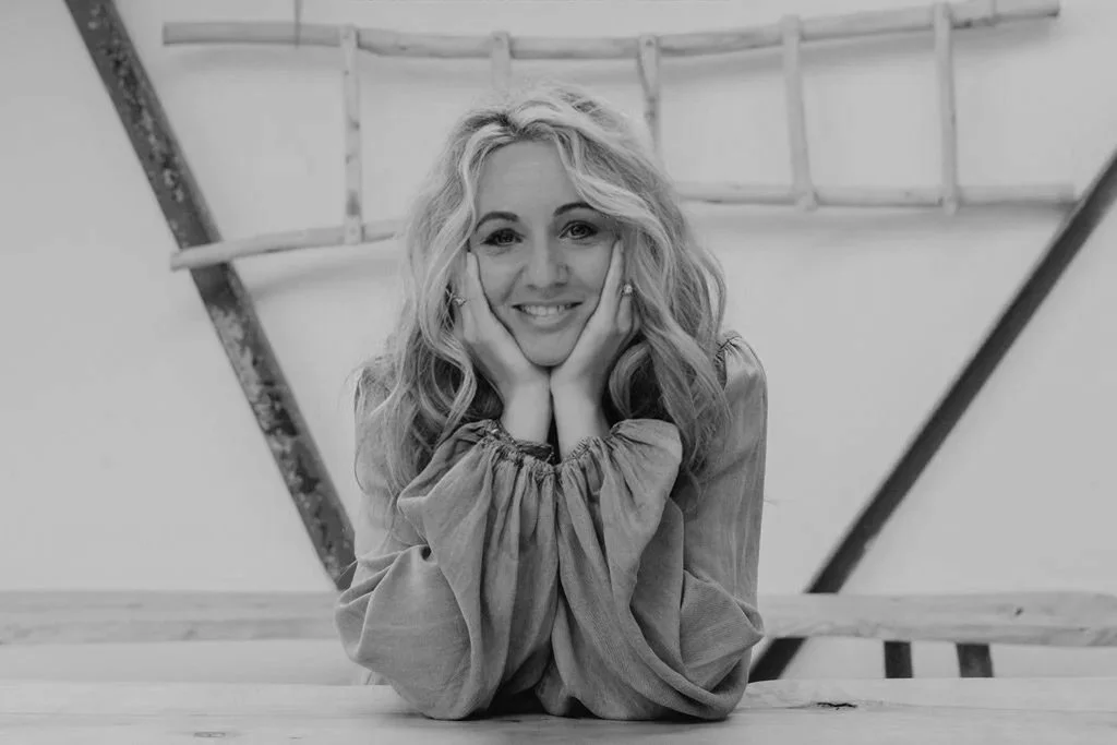 Black and white photo of a woman with wavy hair resting her chin on her hands, smiling at the camera, with a wooden ladder in the background.
