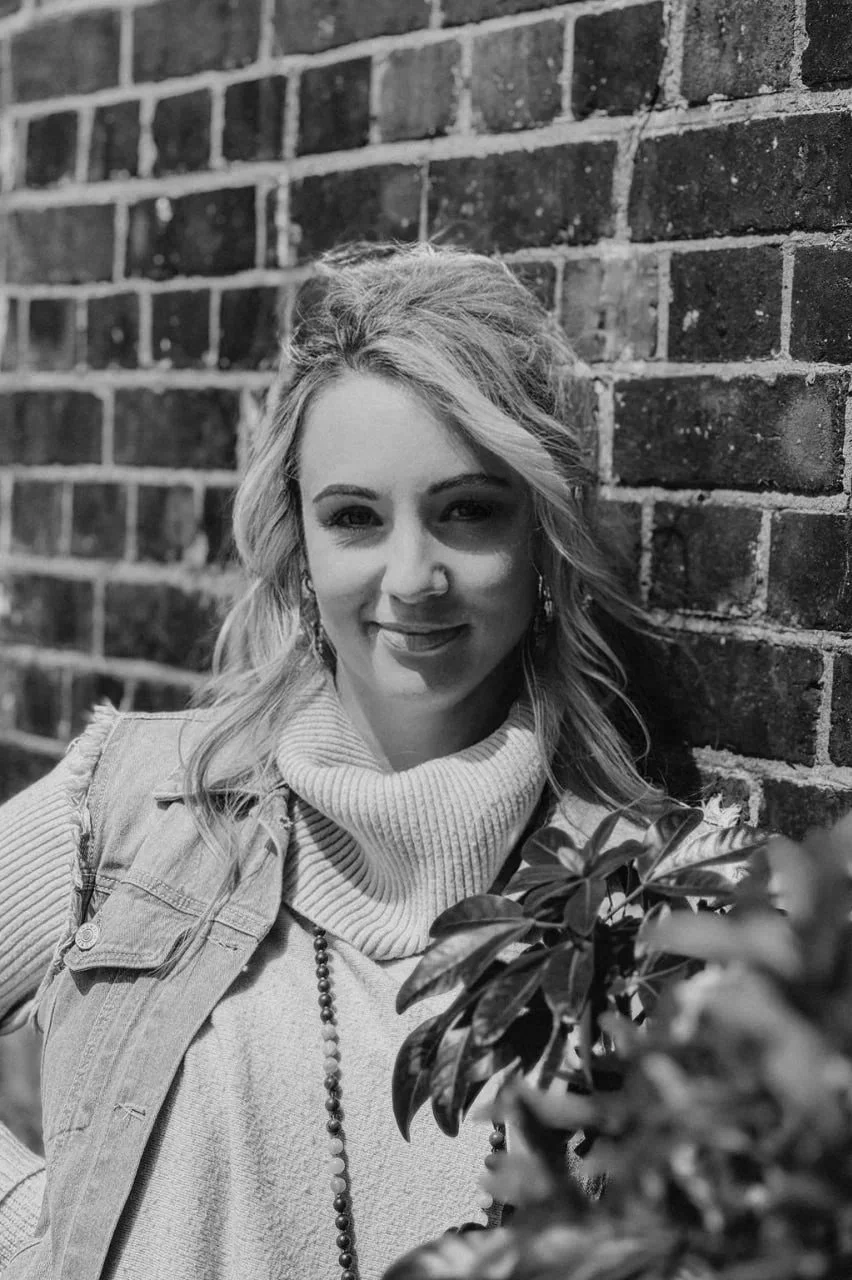A woman with wavy hair leaning against a brick wall, smiling, with a plant in front of her, in black and white.
