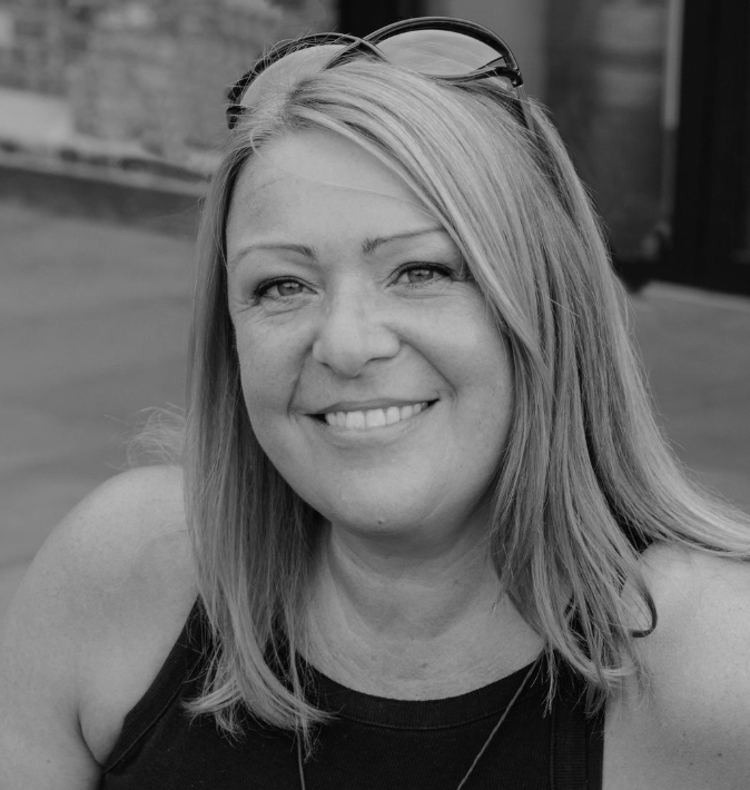 Black and white photo of a smiling woman with shoulder-length hair, wearing sunglasses on her head and a sleeveless top.