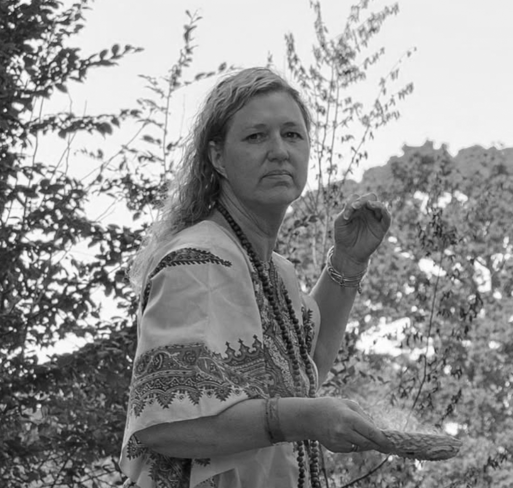 A woman with long, wavy hair standing outdoors in a natural setting with trees and bushes in the background, looking at the camera with a serious expression, holding a object in her right hand, wearing a patterned blouse and multiple necklaces.