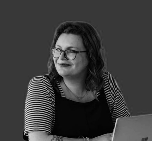 A woman with glasses and wavy hair sitting at a desk with a laptop, wearing a striped shirt and black overalls, smiling and looking to her right.