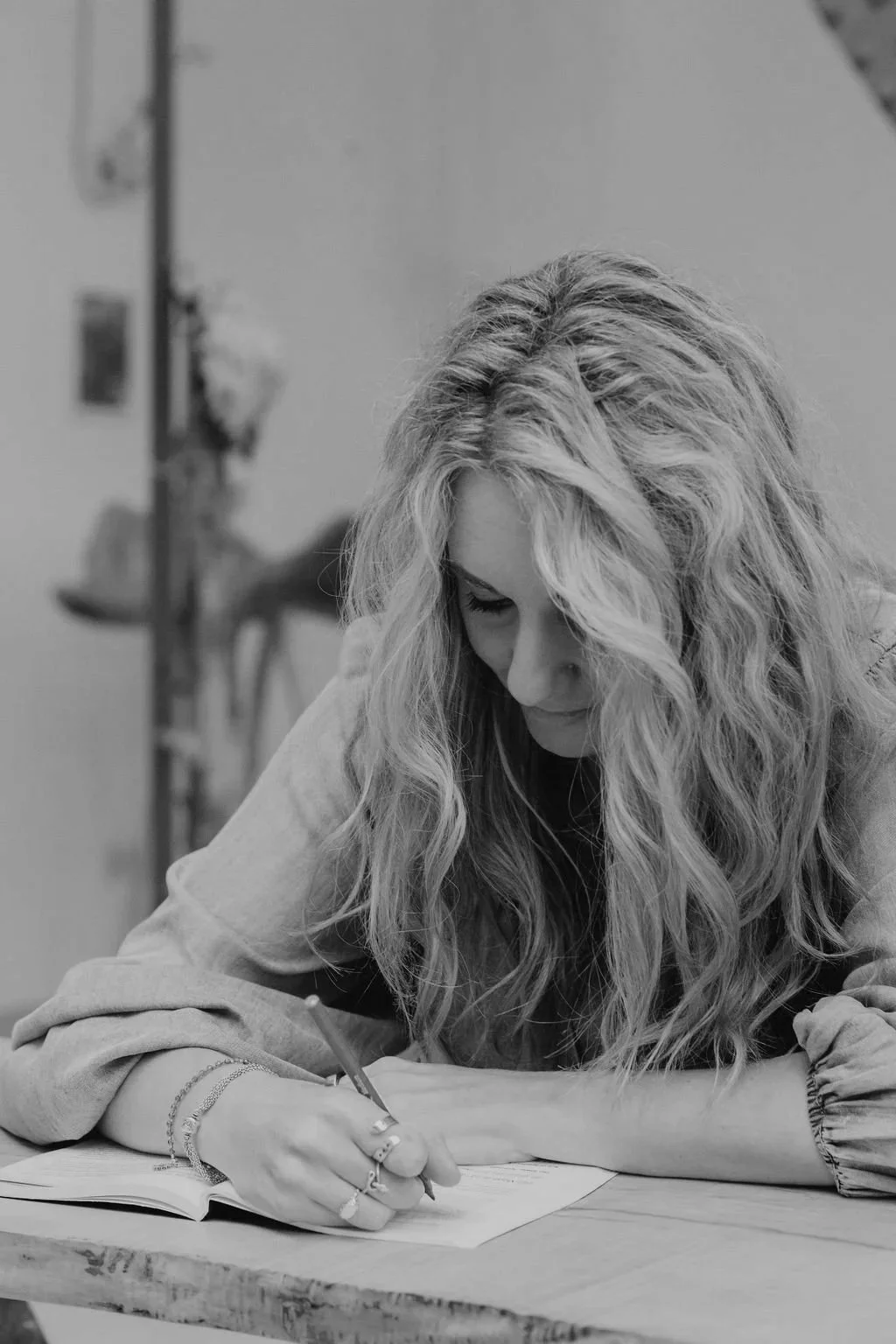 A woman with long, wavy hair writing in a notebook at a desk, in a black-and-white photo.