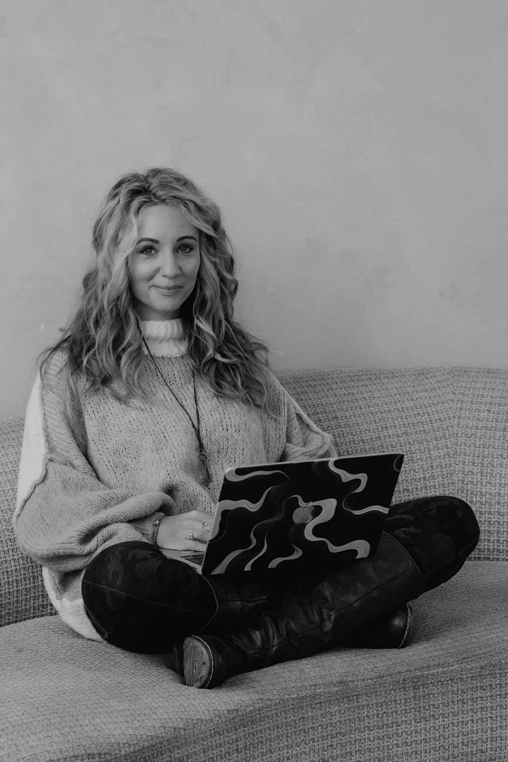 A young woman with curly hair sitting on a couch, holding a laptop with a wavy pattern, smiling at the camera.