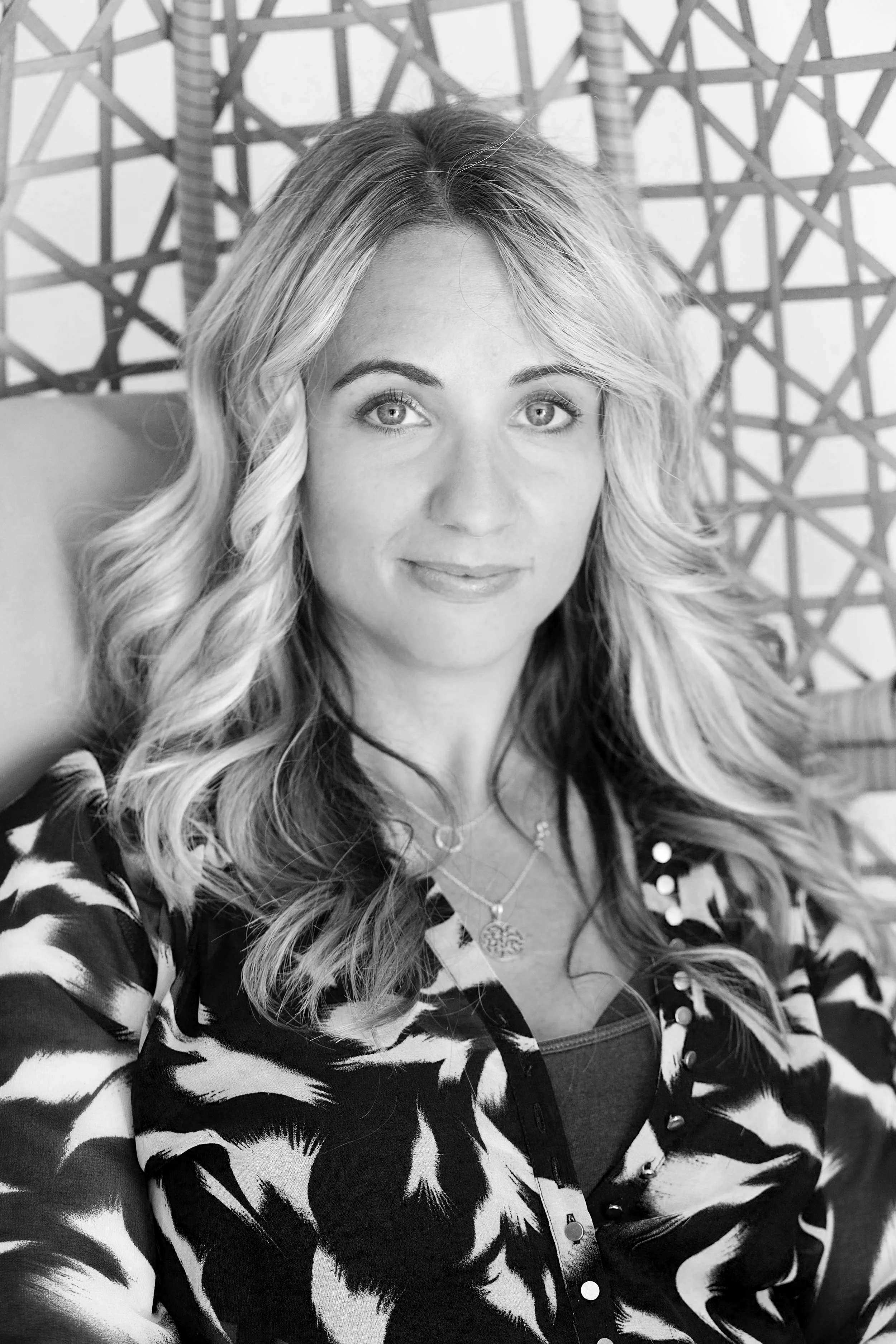 A woman with long curly hair smiling slightly, wearing a patterned jacket and layered necklaces, sitting in front of a woven trellis background.