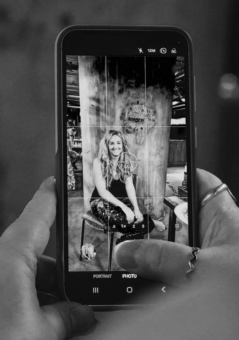 A person taking a black-and-white photo of a woman sitting on a stool in a rustic setting, with a large wooden column and textured wall in the background.