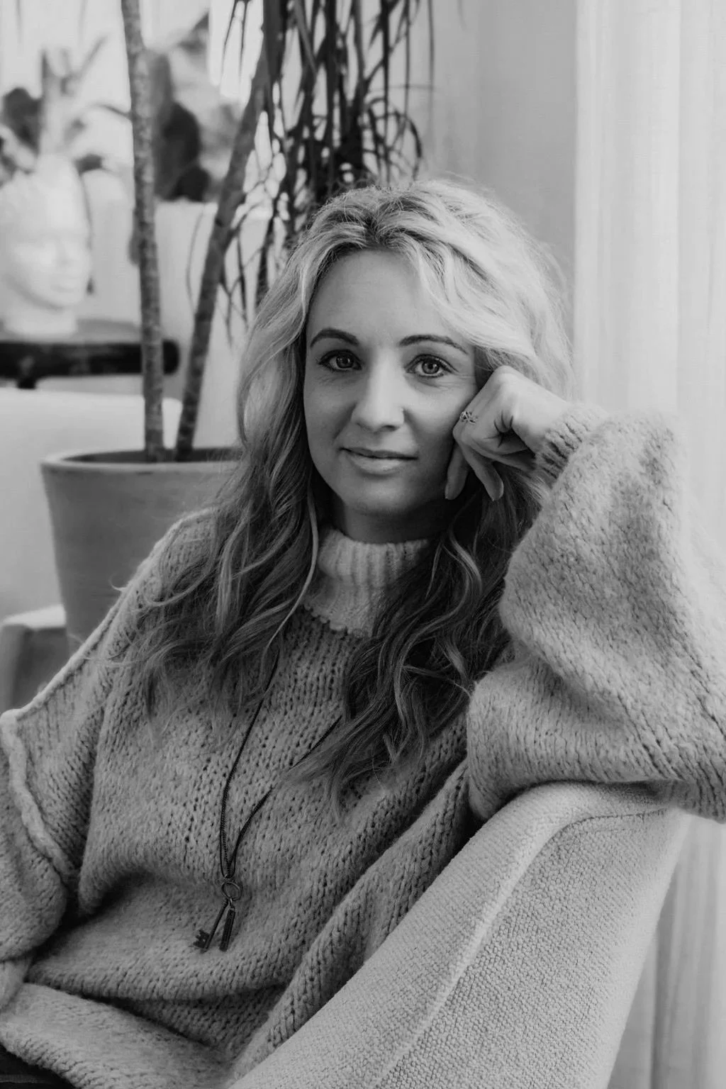 Black and white photo of a young woman with wavy hair sitting in a chair, resting her head on her hand, wearing a thick sweater.