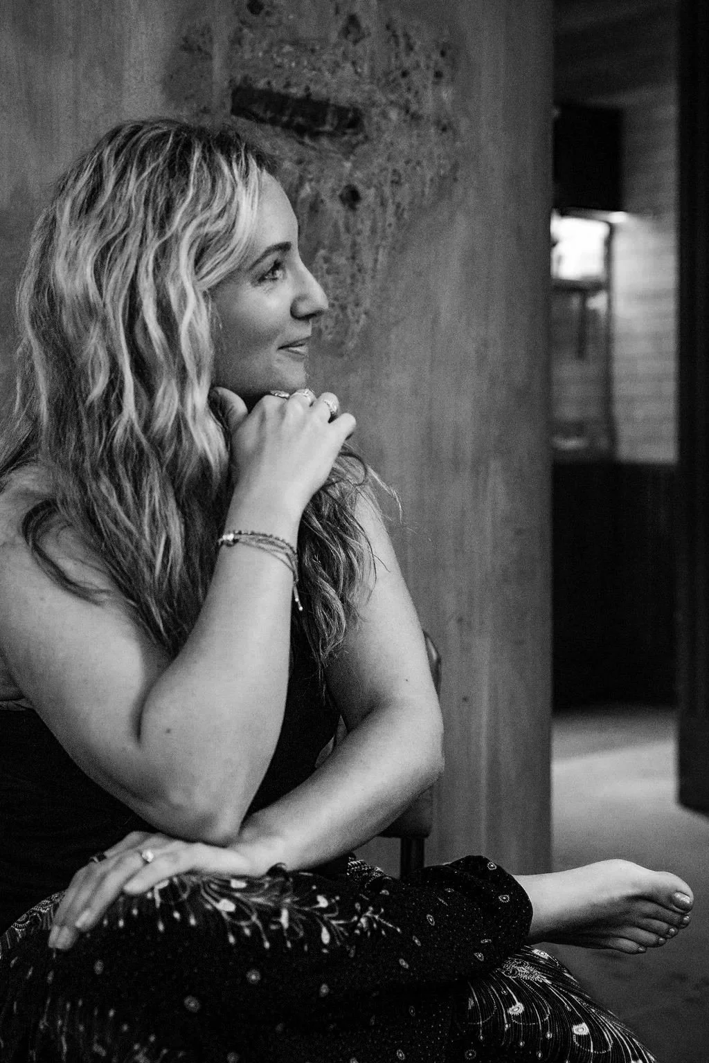 A woman with long, wavy hair sitting with her body turned slightly to the right, resting her chin on her right hand, and looking to the right. She is wearing jewelry, a sleeveless top, and a patterned skirt, sitting indoors near a textured wall.