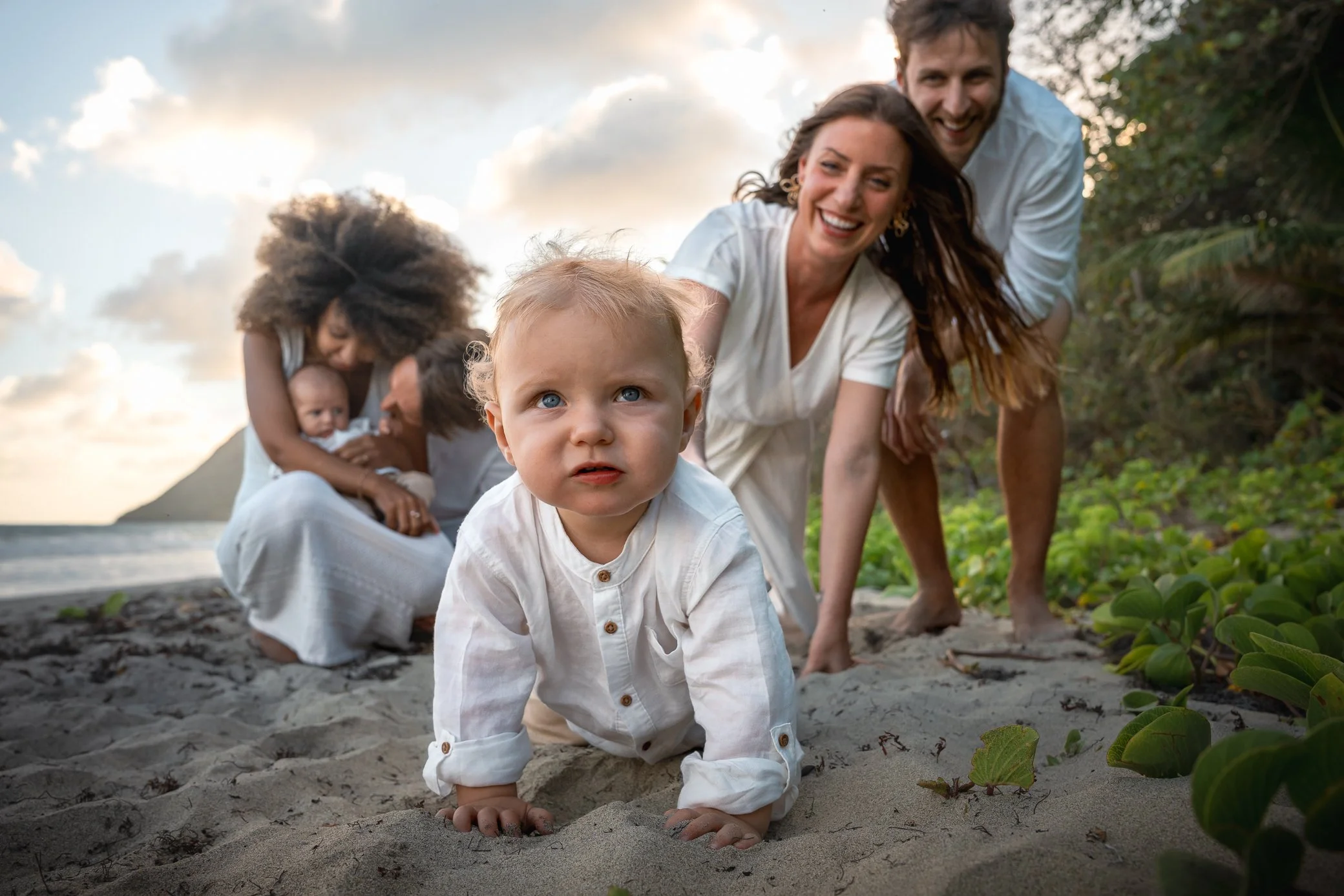 Famille en vêtements blancs sur la plage du Diamant en Martinique, avec un bébé à quatre pattes au premier plan, deux adultes souriants derrière lui et un nourrisson dans les bras d’une femme à gauche, devant la mer et un ciel nuageux.