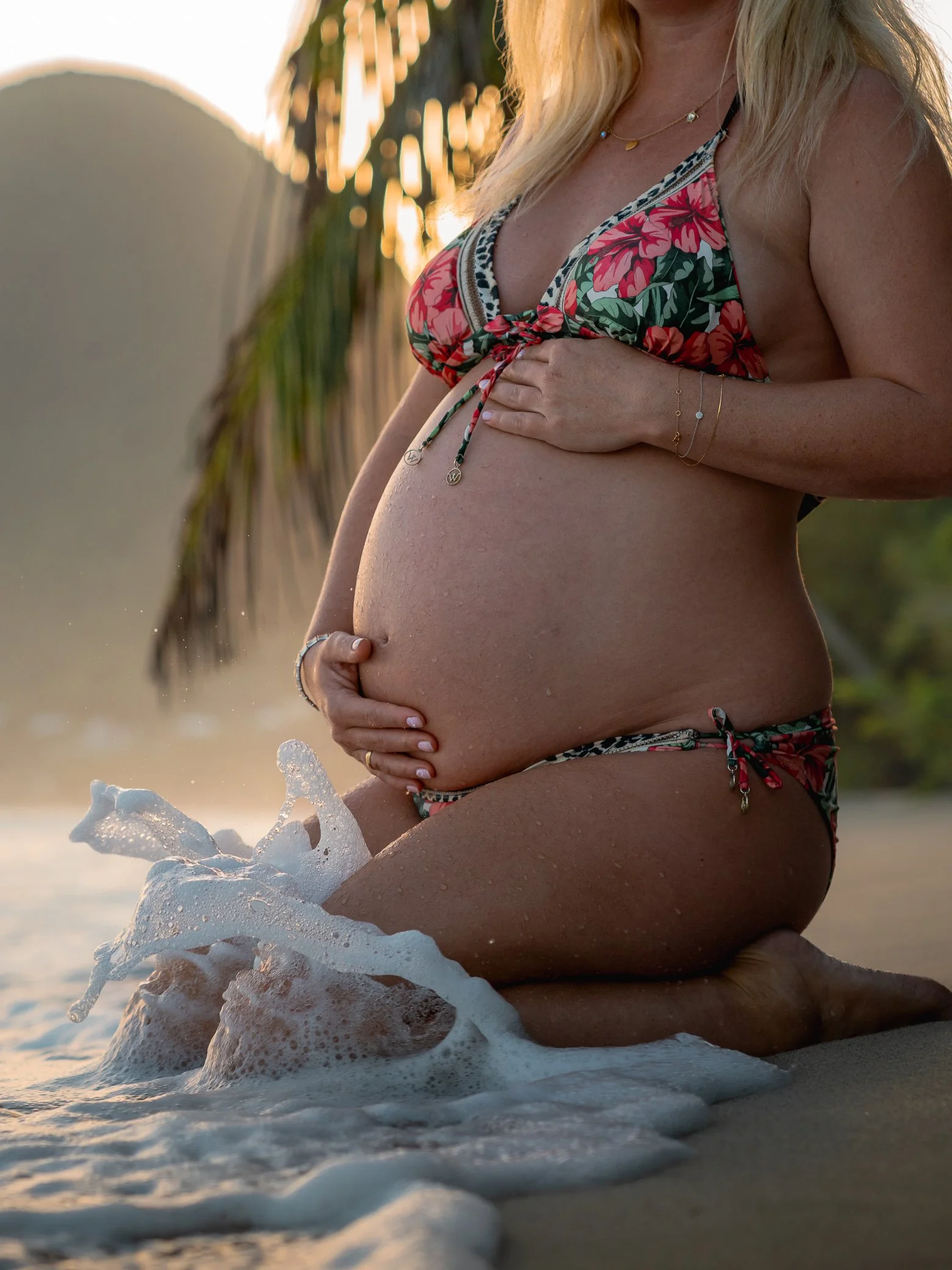 Future maman agenouillée au bord de l’eau lors d’une séance photo de grossesse sur la plage du Diamant de Martinique, les mains sur le ventre, une vague qui éclabousse au premier plan dans la lumière dorée