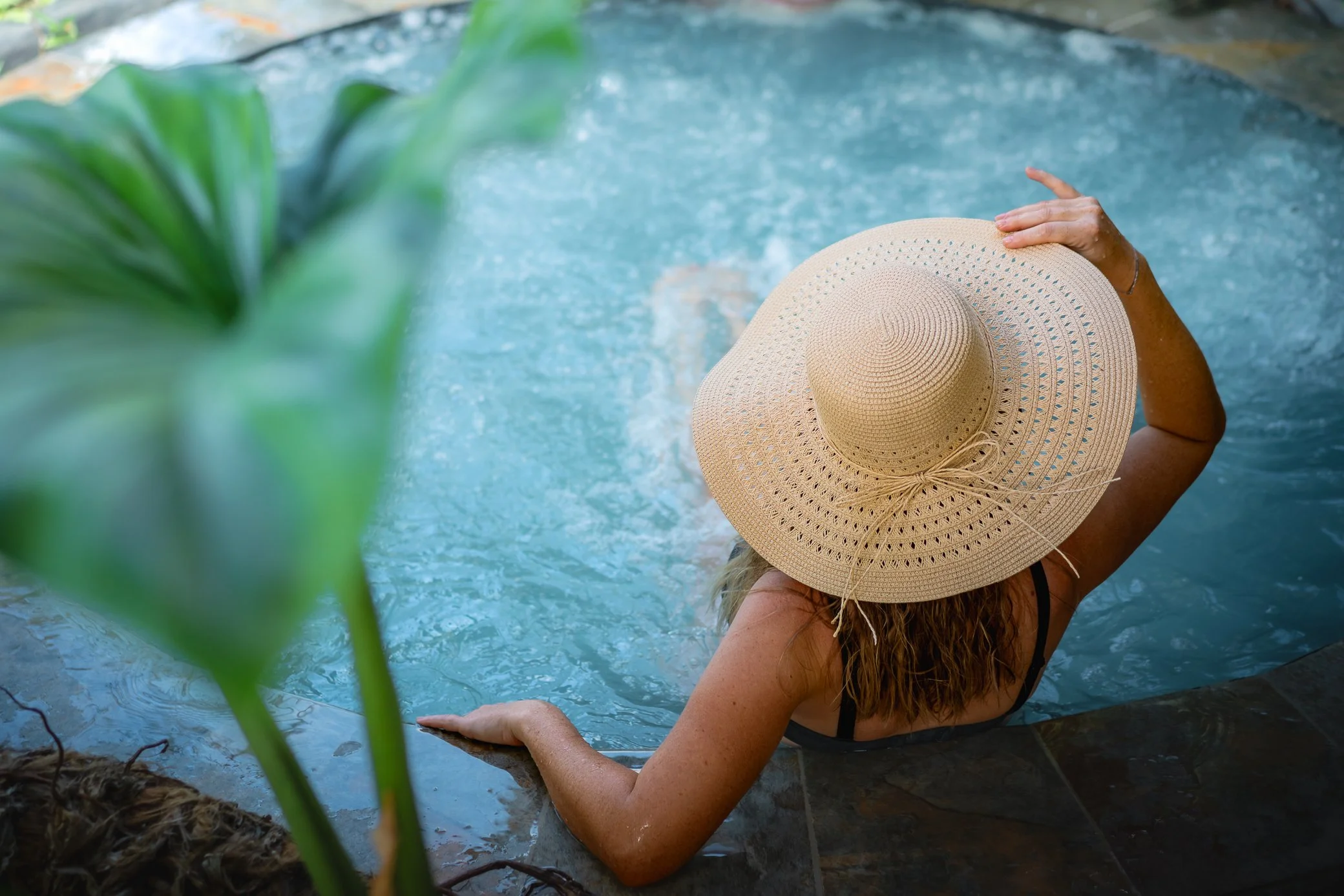 Femme de dos, coiffée d’un chapeau de paille, assise au bord d’un bassin d’eau turquoise en Martinique, avec une grande feuille verte au premier plan.
