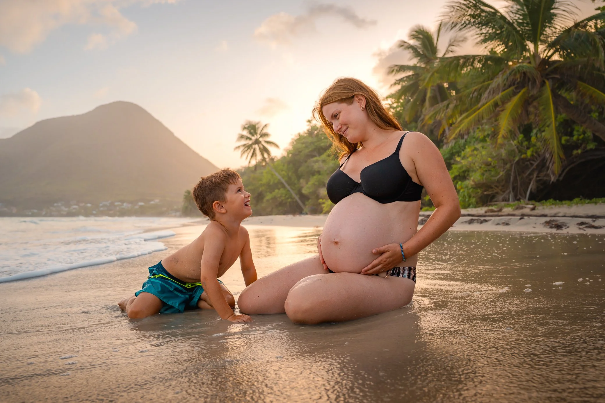 Une femme enceinte assise sur le sable mouillé échange un sourire avec son jeune fils, sur une plage de Martinique au coucher du soleil