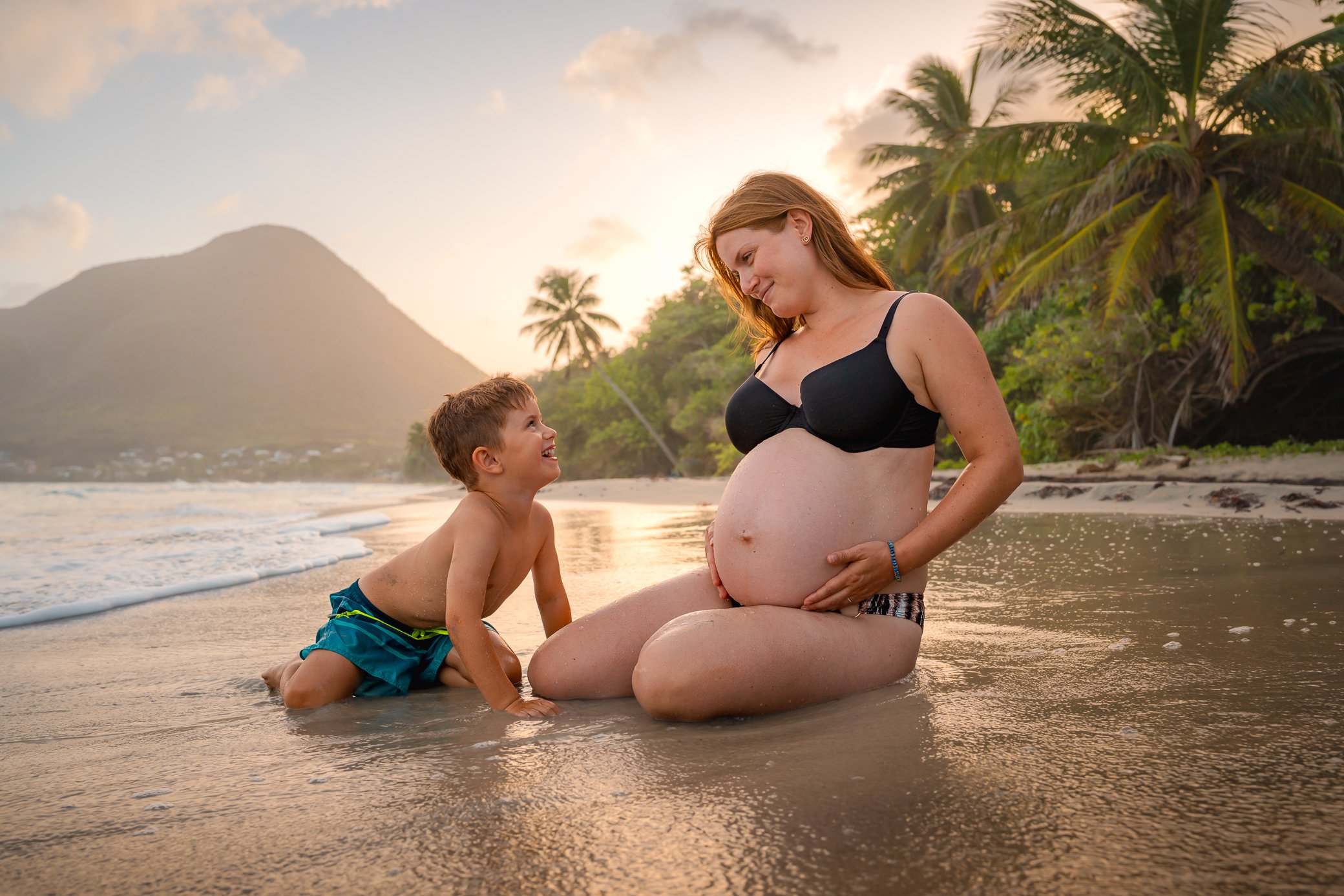 Femme enceinte assise au bord de l’eau sur la plage du Diamant en Martinique, son fils agenouillé à côté d’elle la regarde