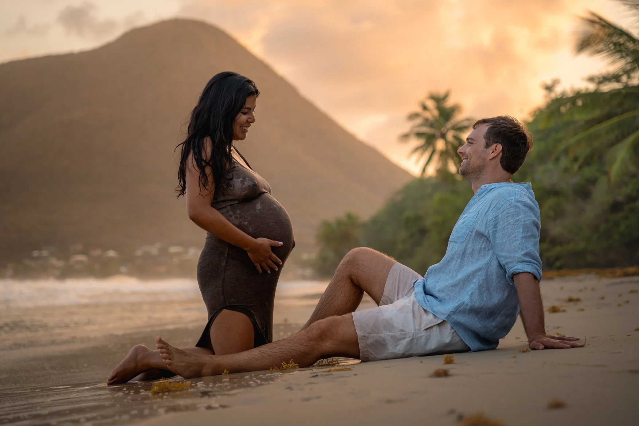 Une femme enceinte à genoux sur la plage sourit en regardant son ventre, un homme assis près d’elle la regarde, devant une montagne au coucher du soleil