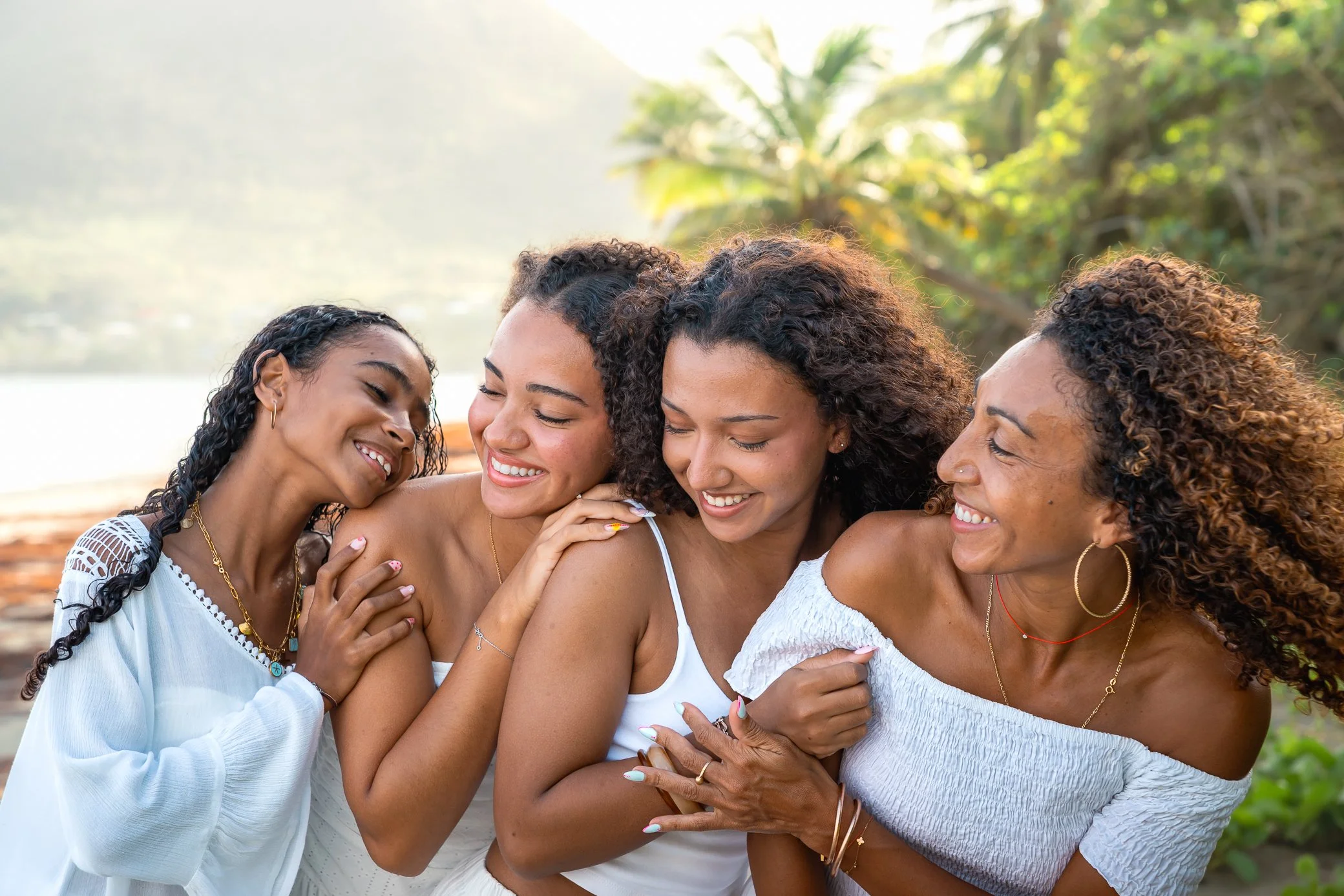 Une mère et ses trois filles, vêtues de blanc, enlacées et souriantes lors d’une séance photo de famille sur la plage du Diamant en Martinique, avec des palmiers derrière elles.