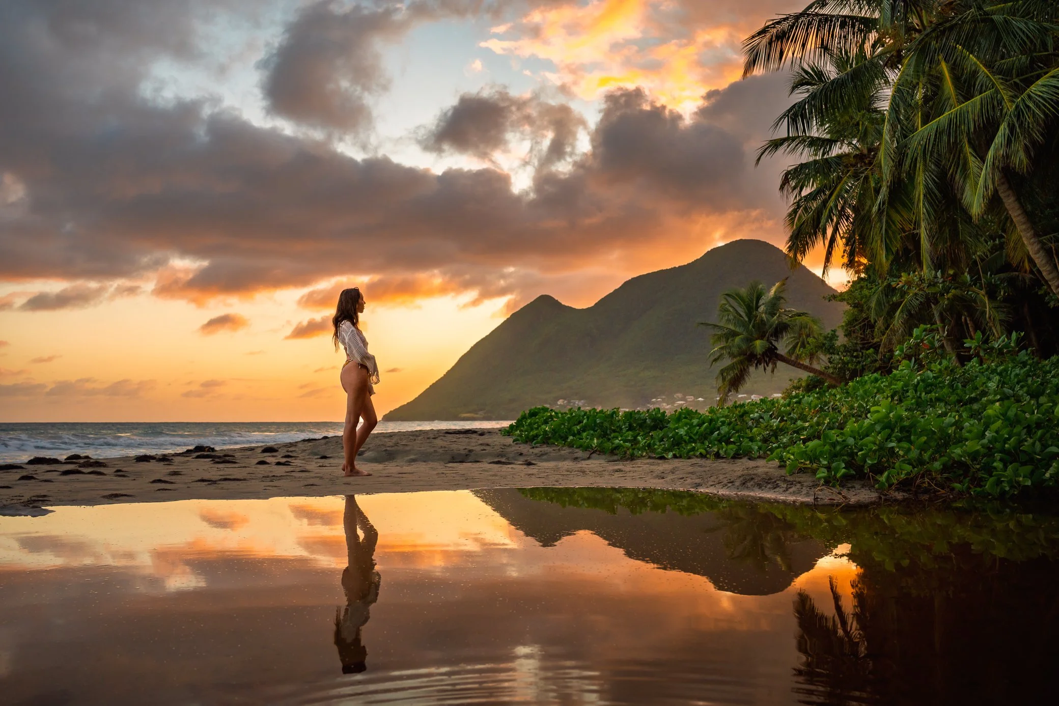 Femme debout sur la plage du Diamant au coucher du soleil, avec une montagne et des palmiers en arrière-plan, et leur reflet du morne Larcher dans l’eau au premier plan.