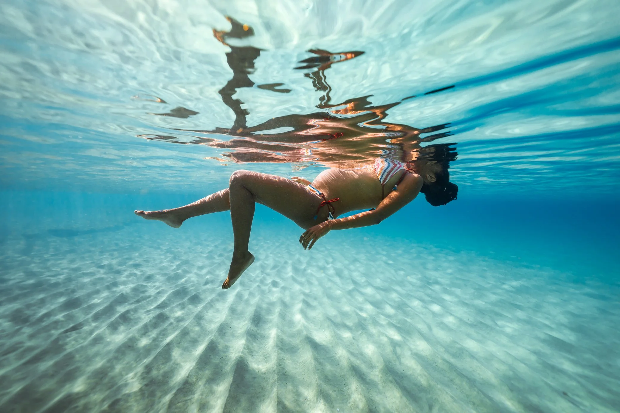 Une femme enceinte flotte à la surface, vue de dessous, dans une eau turquoise limpide en Martinique