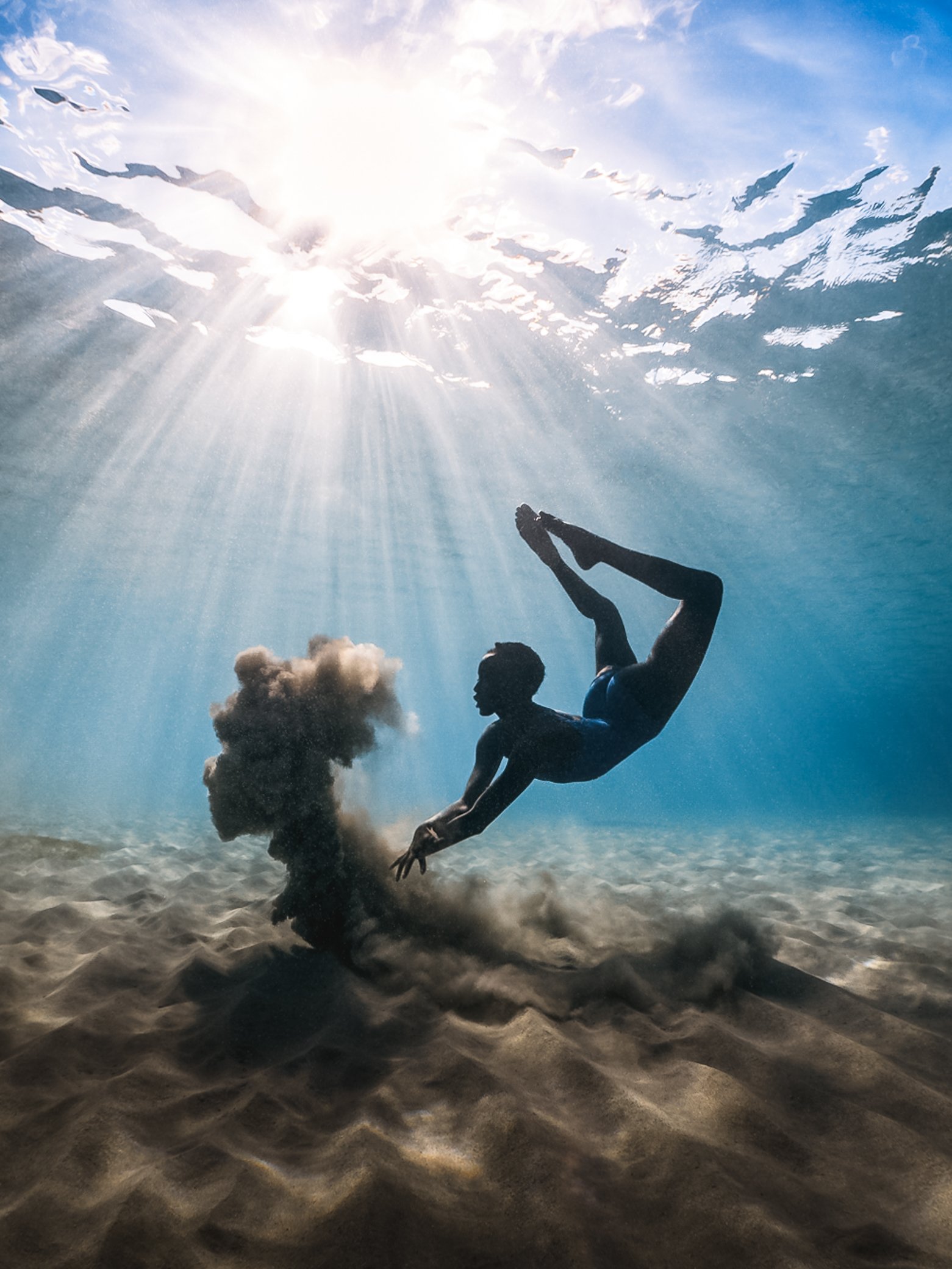 Un e apnéiste descend tête en bas vers le fond sableux, dans une eau claire traversée de rayons de soleil