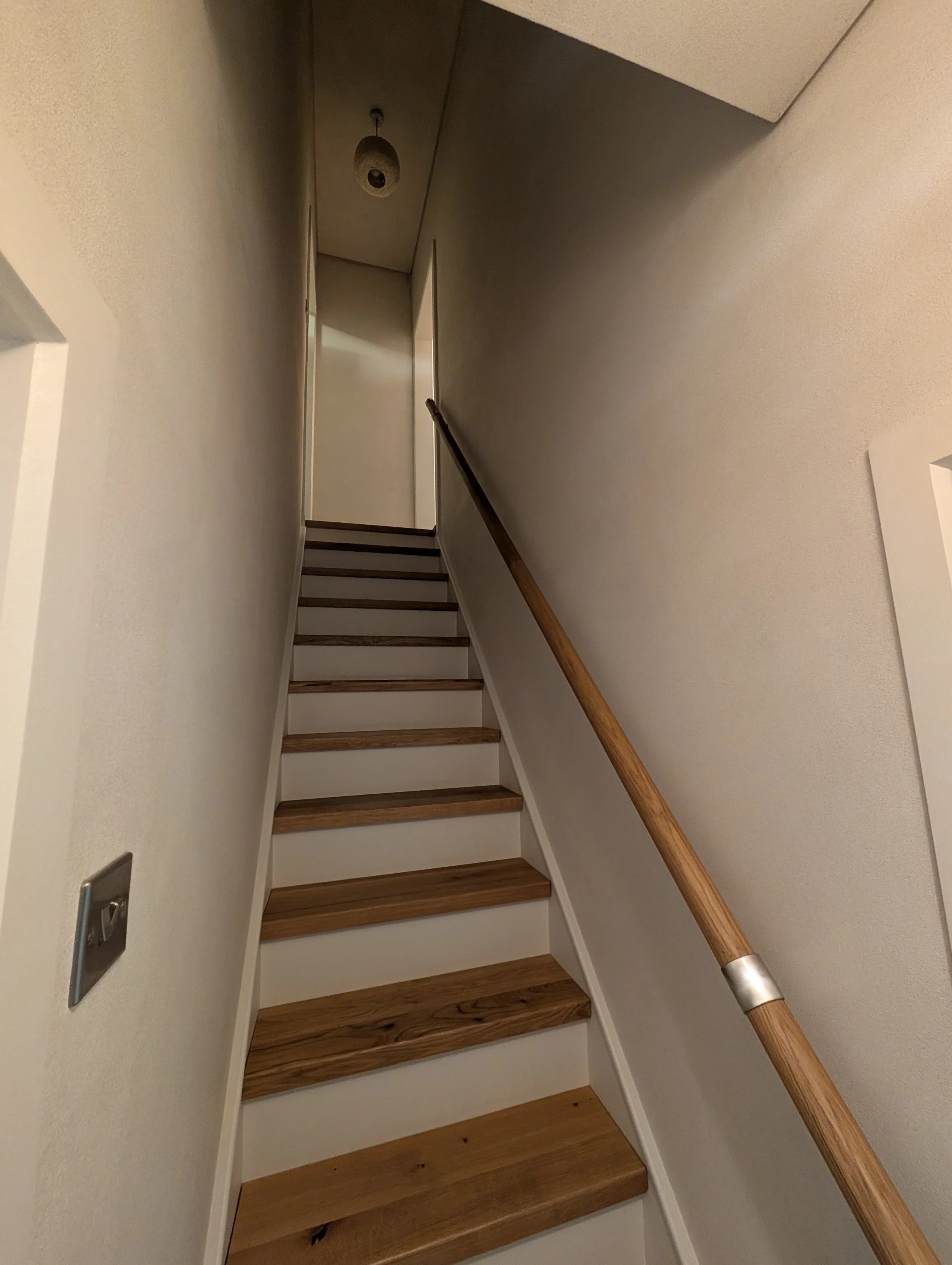 Interior view of a staircase leading upstairs with a wood handrail on the right side, beige walls, a ceiling light fixture, and a wall switch on the left.