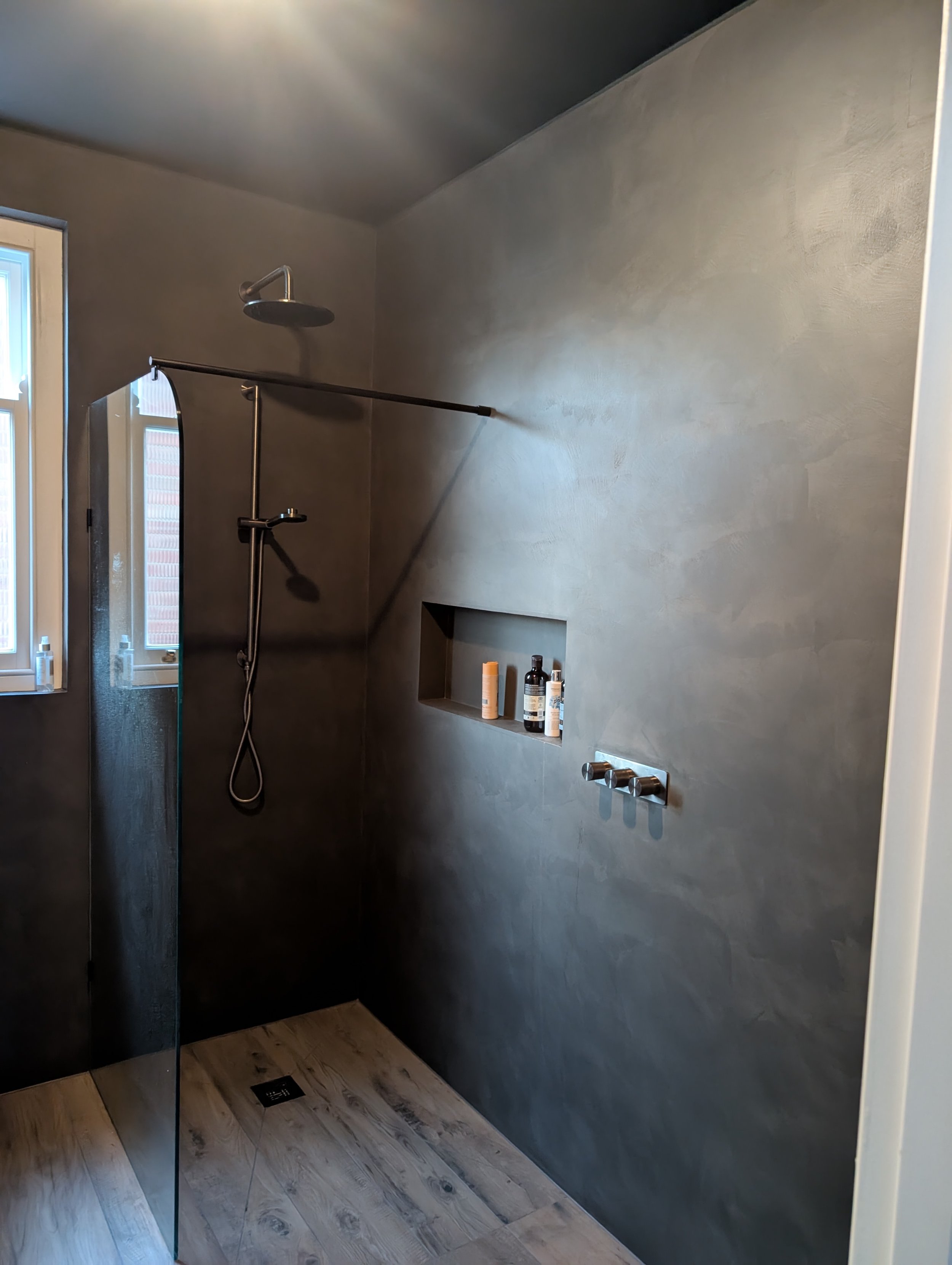 Modern bathroom with a black wall, a rainfall showerhead, a handheld shower, built-in wall niche with toiletries, a window, and wooden floor tiles.