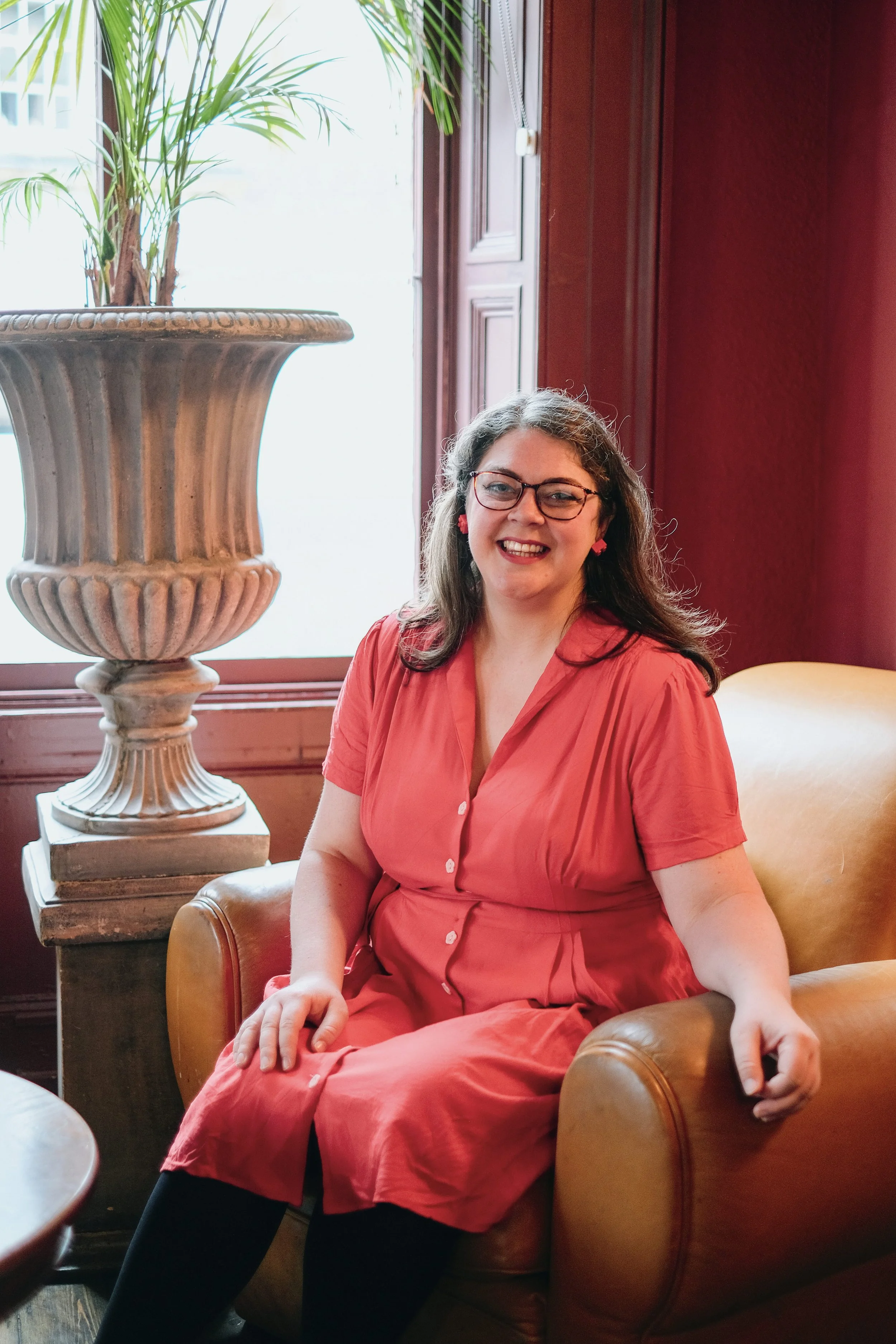 Laura, a woman wearing a red dress sits in a brown armchair with a plant behind her