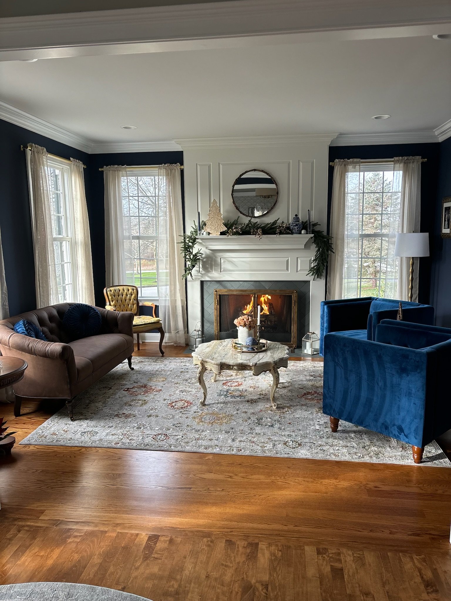 Interior view of a living room corner featuring a white fireplace with black insert, a tall dark blue built-in cabinet above the fireplace, three large windows with grid panes, and some photos and decorative items on the mantel.