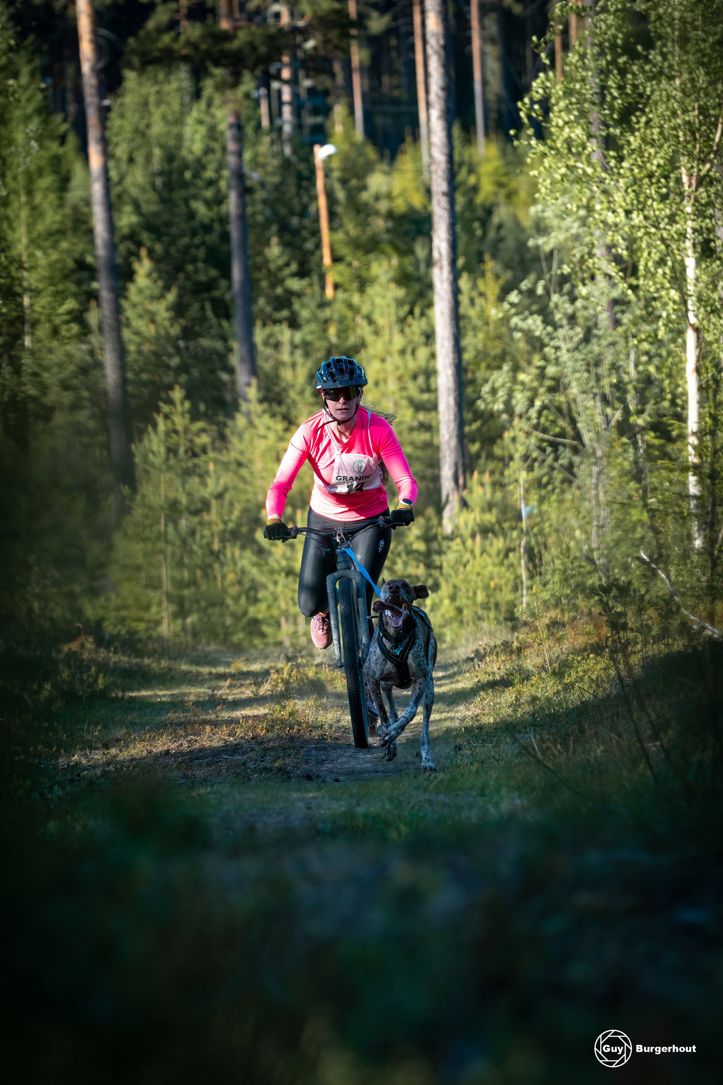 A woman riding a mountain bike with a dog in a forest trail during daytime. The woman is wearing a pink long-sleeve shirt, helmet, and sunglasses, with a race bib labeled 'GRANIN'. The forest has tall trees and dense green foliage.