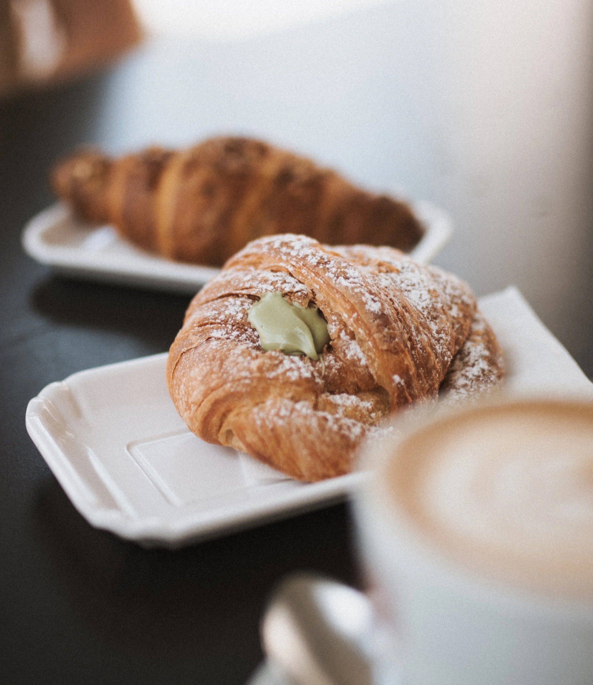 Close-up of a pistachio-filled cornetto with another pastry and a cappuccino in the foreground