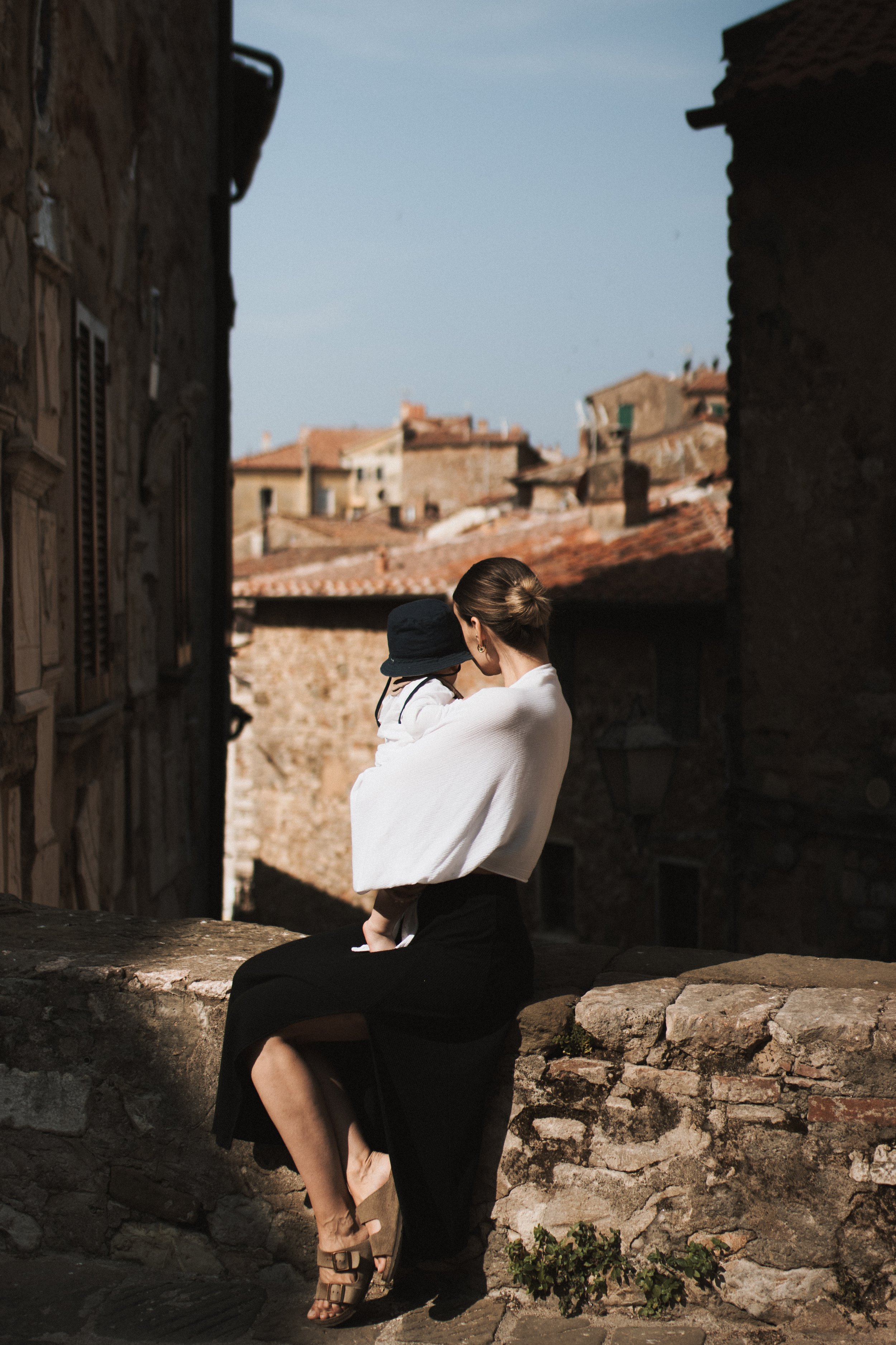 Mother holding her toddler on a stone wall, looking through a gap between stone walls over the rooftops of Campiglia Marittima