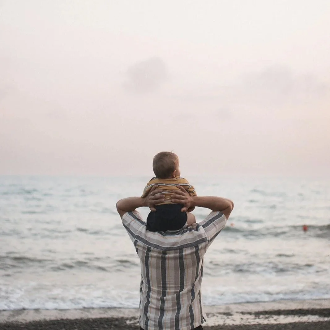 Father carrying his toddler on his shoulders as they watch the sunset over the sea on the Tuscan coast