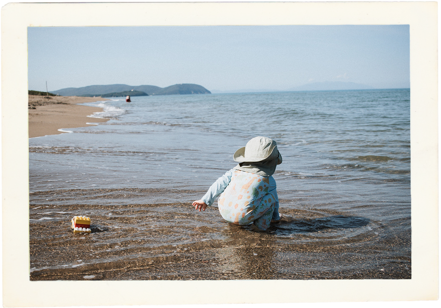 Toddler playing in the shallow sea at Spiaggia di Rimigliano, with the sandy coast and Tuscan hills in the distance