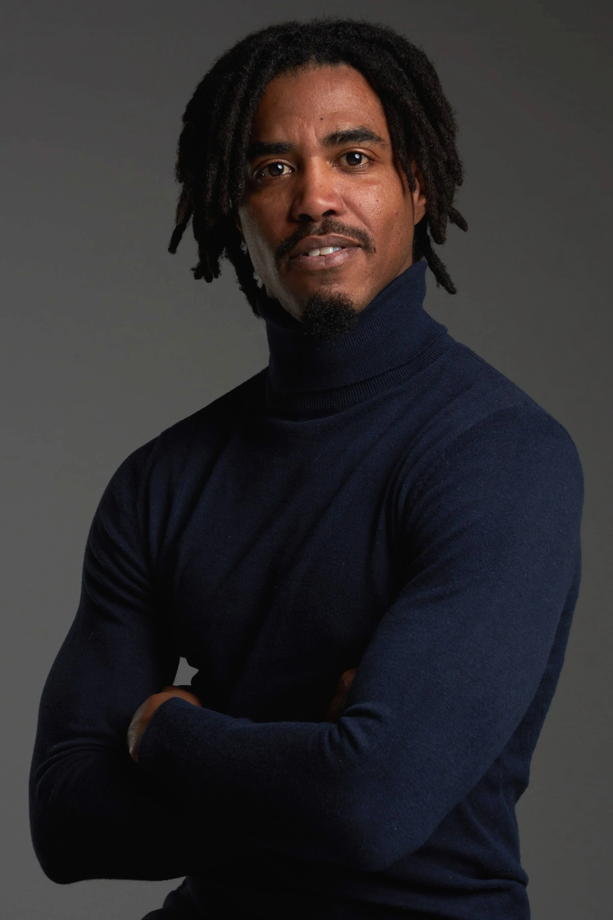 Hombre con cabello rizado y barba, usando suéter de cuello alto azul marino, cruzado de brazos frente a un fondo gris oscuro.