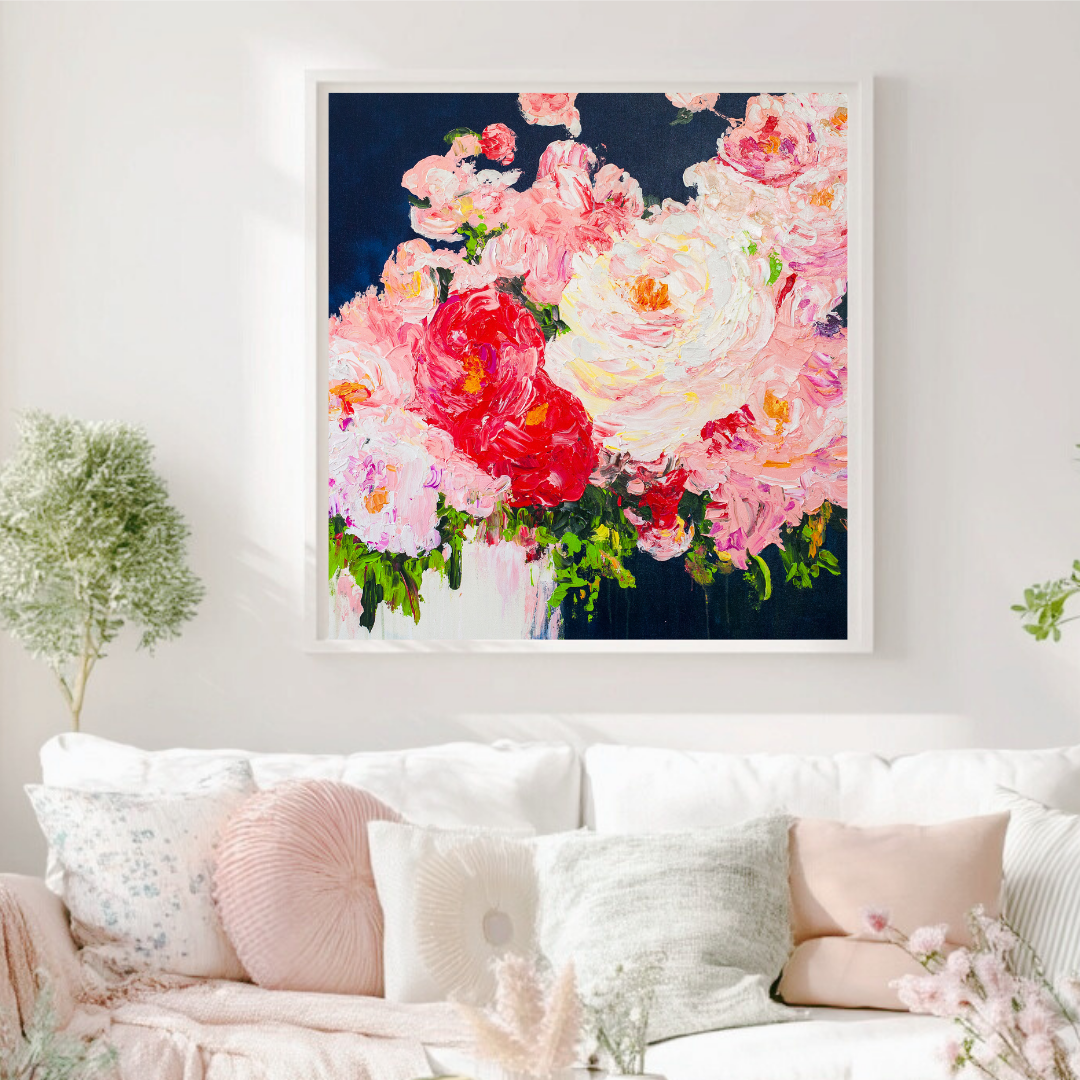 A vibrant abstract flower painting with pink, red, and white flowers on a dark background, hung above a white sofa decorated with pastel-colored pillows and pink flowers, in a modern living room.