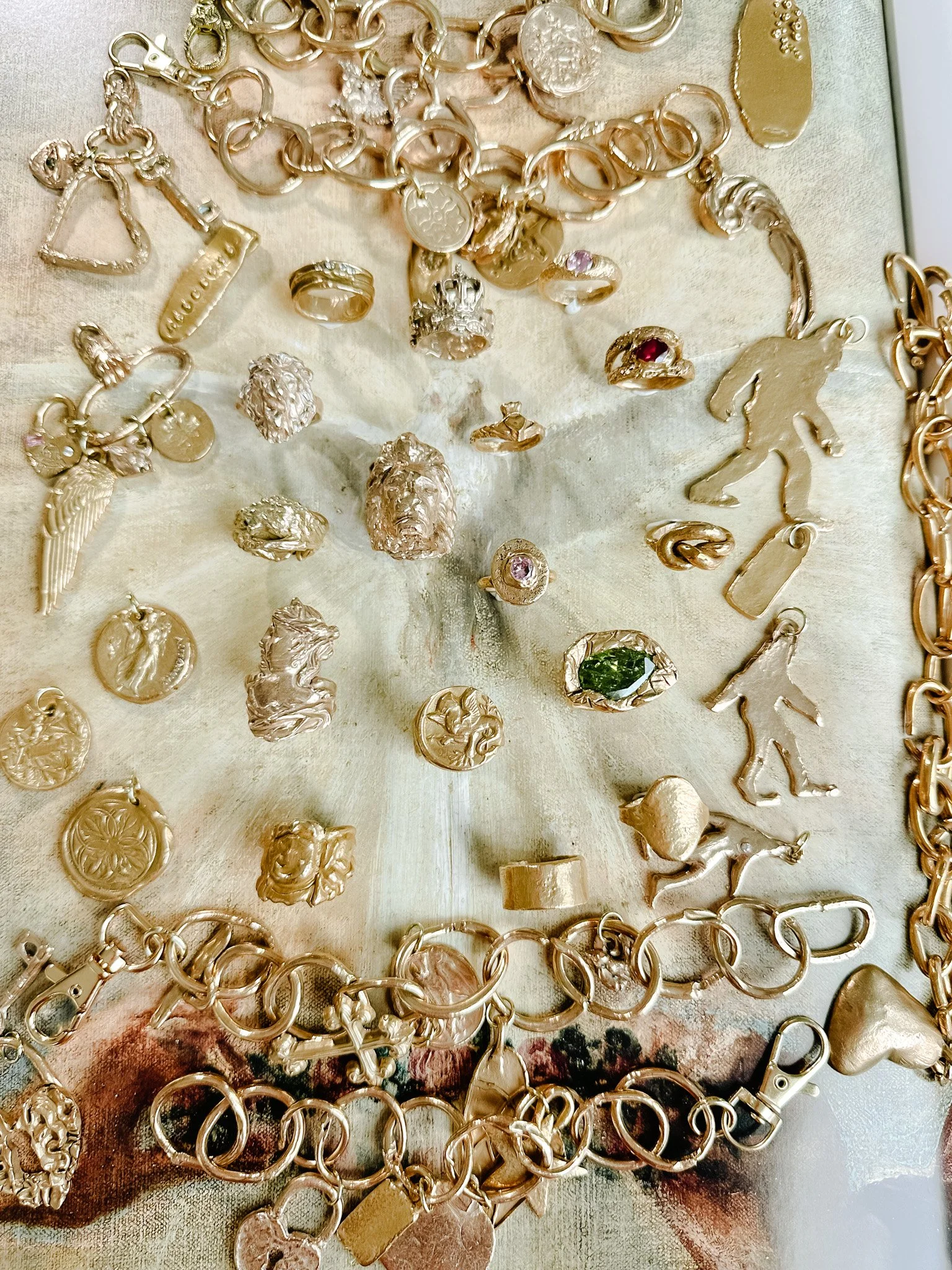 A collection of various gold jewelry pieces including necklaces, bracelets, and rings, arranged on a light-colored surface. The jewelry features different charms, Medallions, and designs like lions, angels, and abstract shapes.