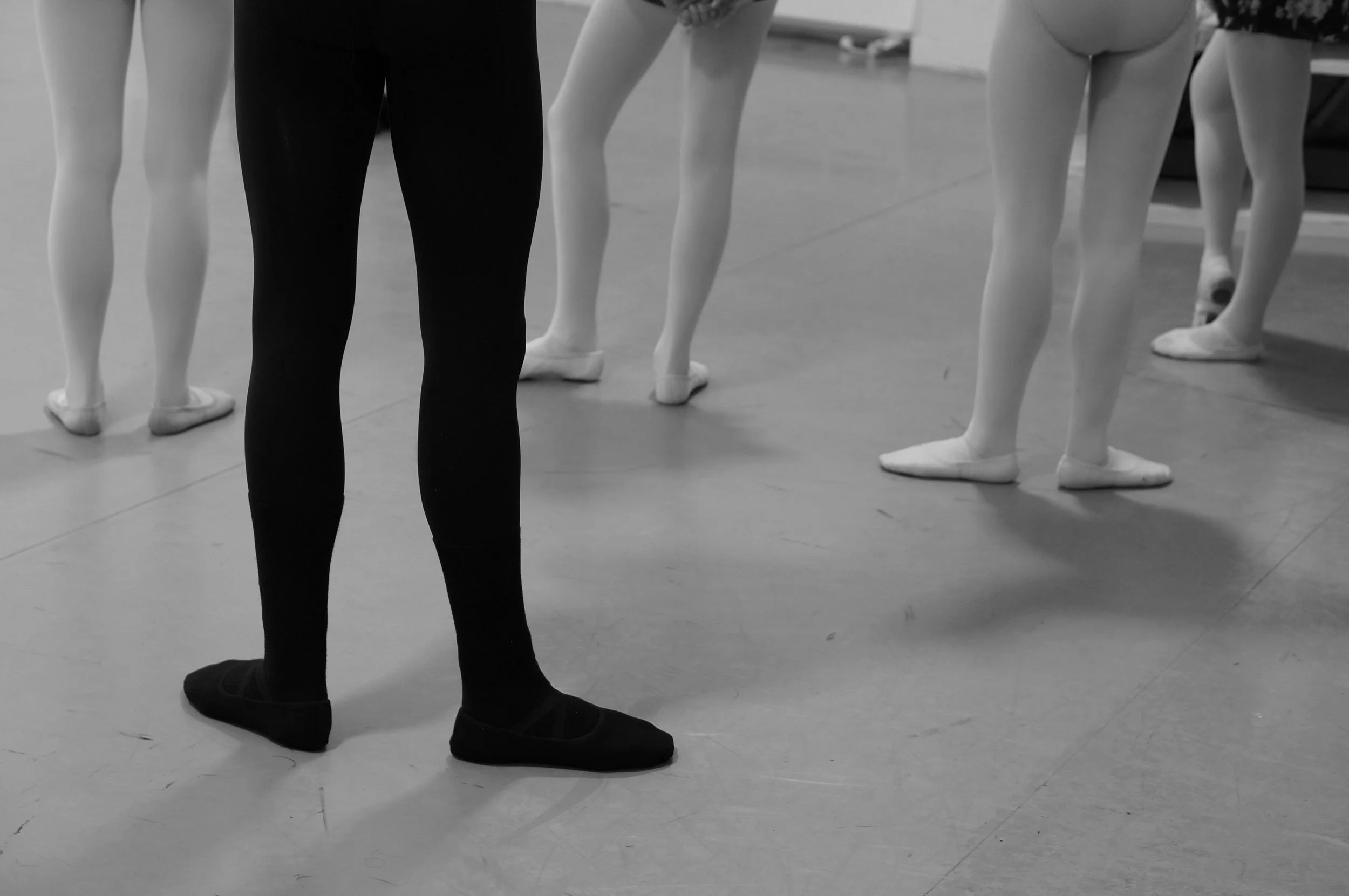 Ballet dancers in rehearsal, with a focus on their legs and feet, and a dance instructor in black tights observing.