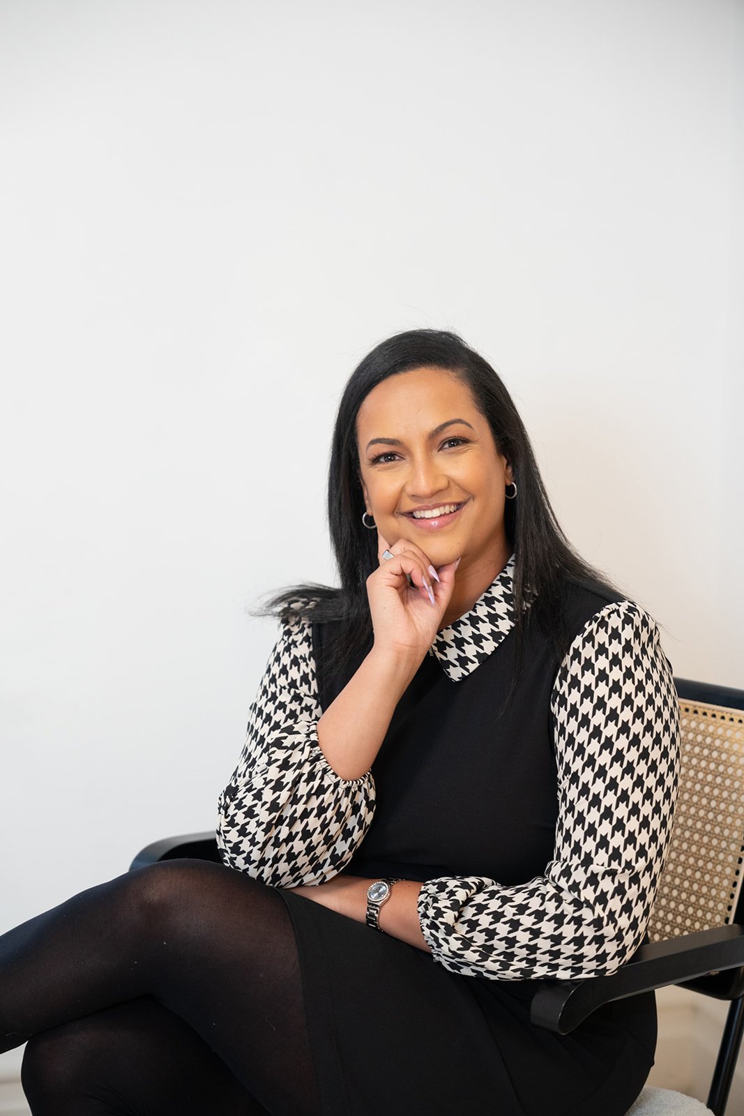 A woman with dark hair, smiling, seated on a chair, wearing a black dress and houndstooth patterned blouse.