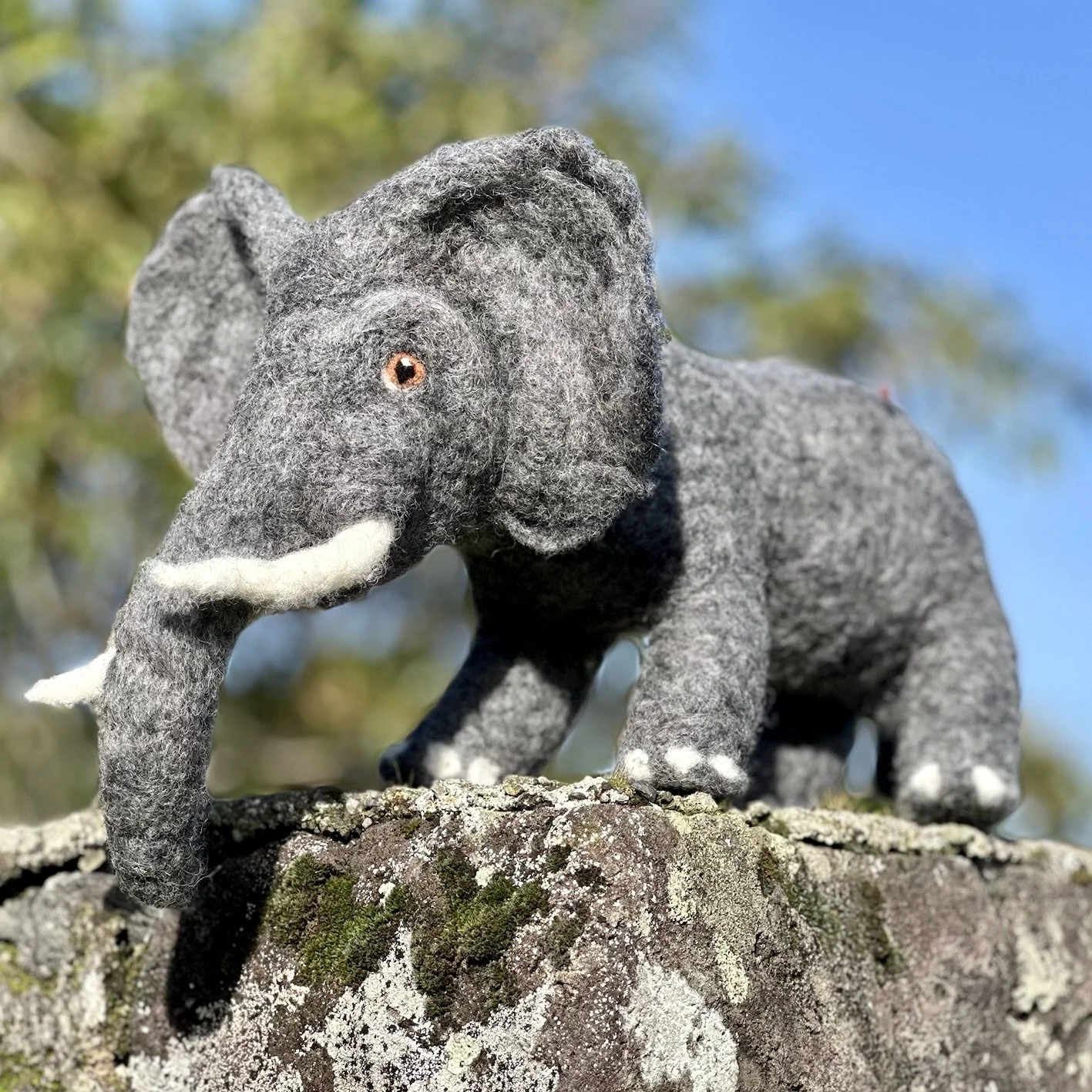 A plush gray elephant toy with floppy ears, white tusks, and orange eyes, standing on a moss-covered log with a blue sky and blurred trees in the background.