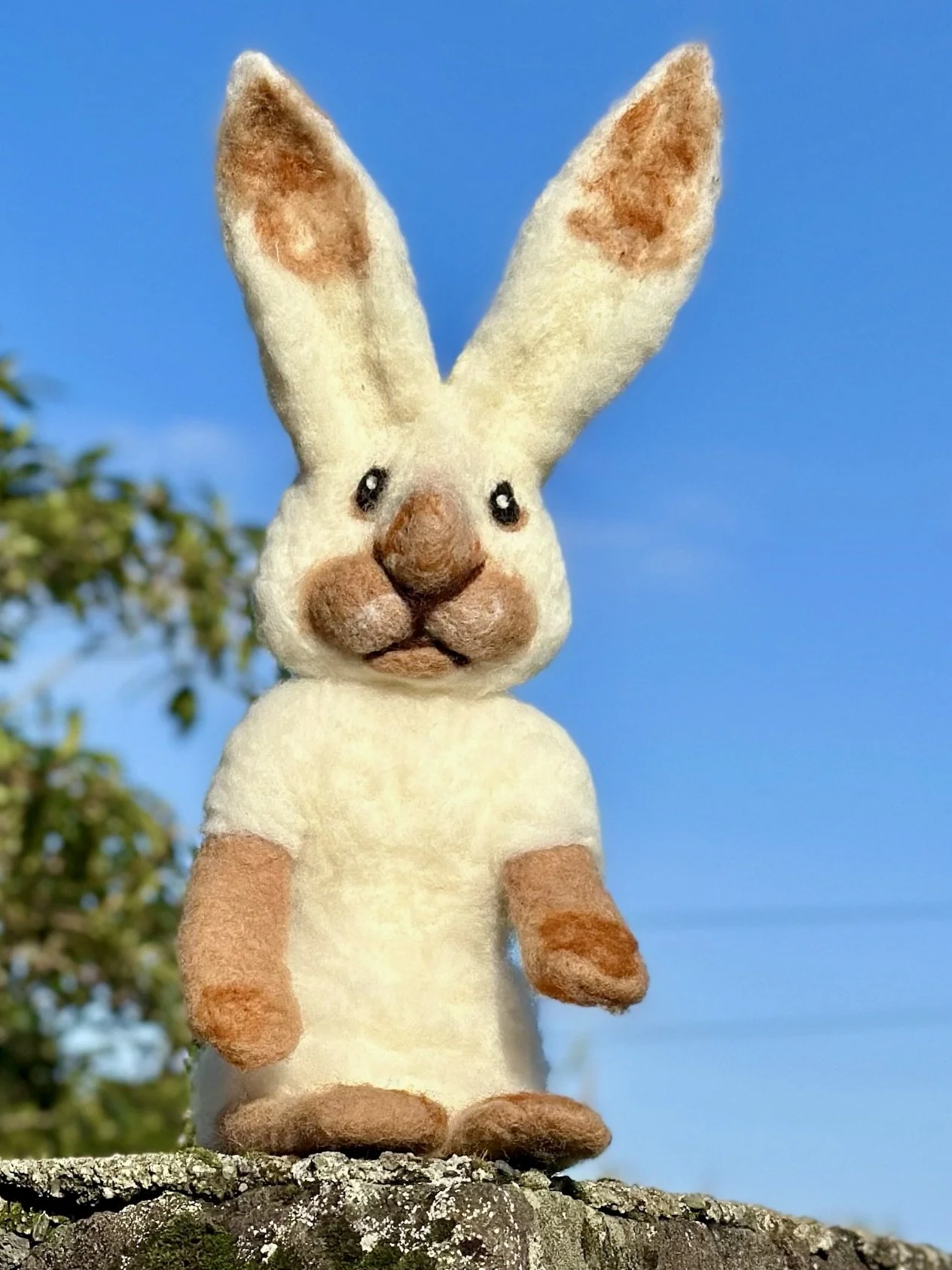 A plush toy of a bunny with tall ears, standing outdoors on a rock, with a blue sky and some tree branches in the background.